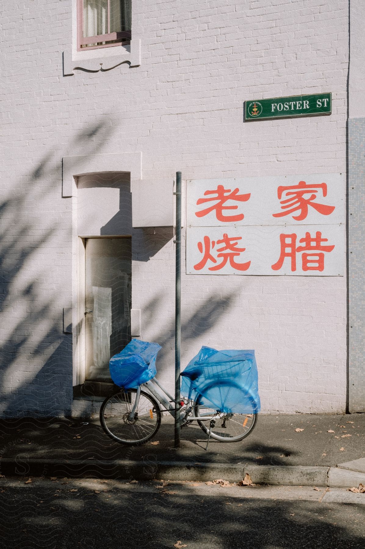 A bicycle is parked next to a building with a blue tire cover. A sign that says "Foster Street" is above the bicycle, with some Chinese characters written in red below it.