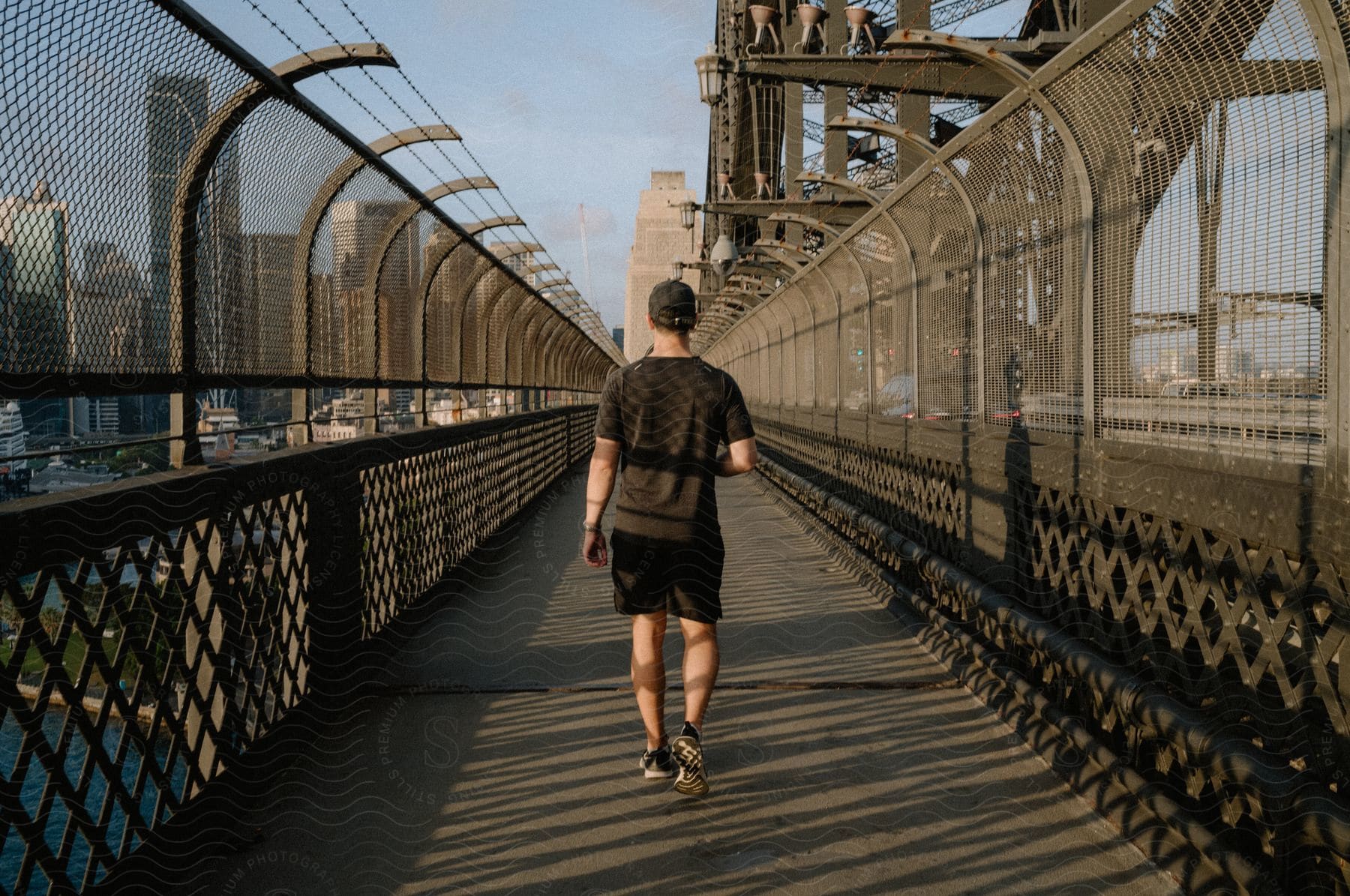 A man wearing a cap, T-shirt, and black shorts walks across the Sydney Harbour Bridge.