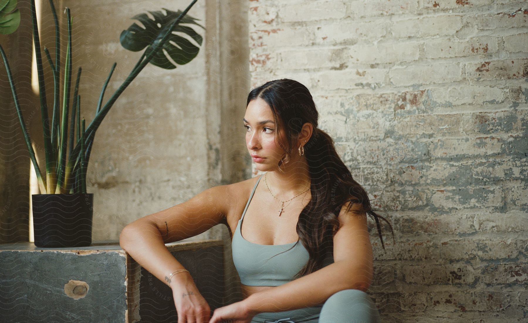 A woman with long dark hair is sitting by a wall with her elbow resting near a potted plant as she looks to her side