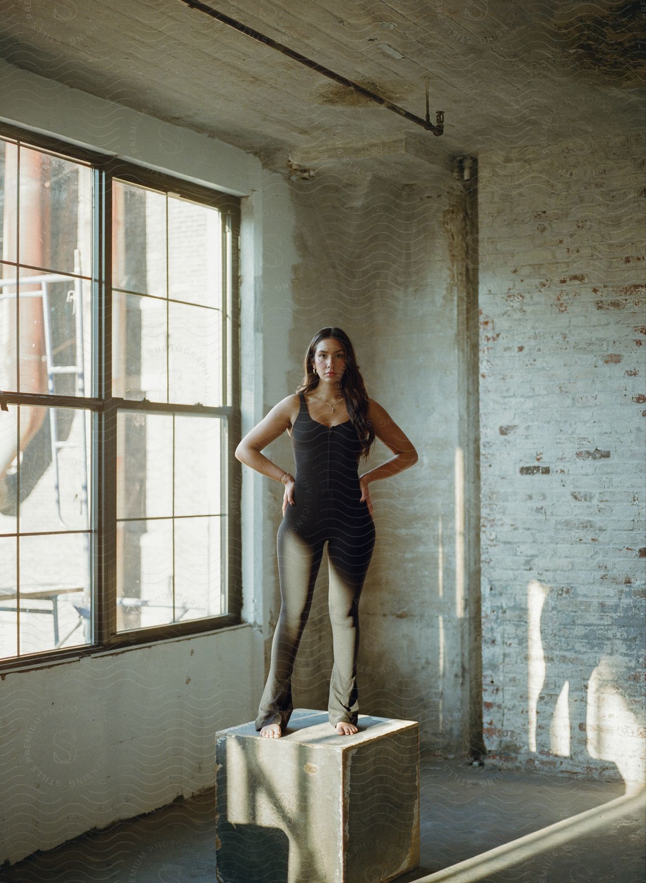 Woman in a black outfit standing on a pedestal in a sunlit industrial space with large window and white brick wall.