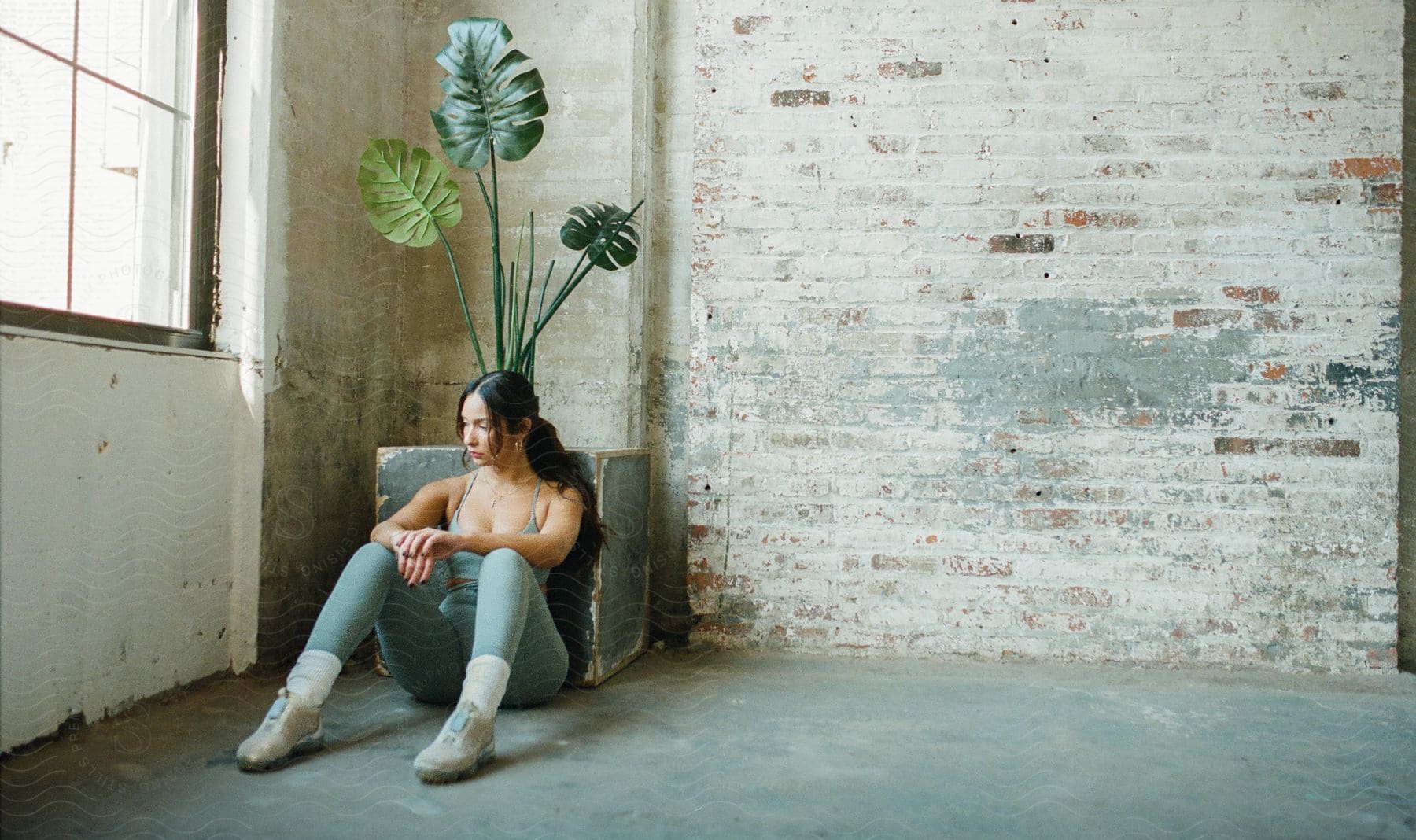 A woman with long dark hair is sitting by a wall with her elbow resting near a potted plant as she looks to her side