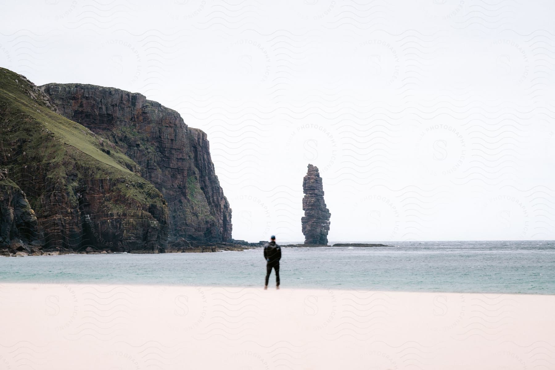 Man with his back to the camera on the beach with the sea coastline and geological formations beside him.
