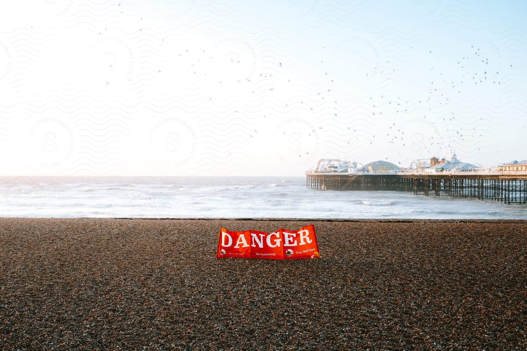 A danger sign on a beach next to a pier covered in buildings.