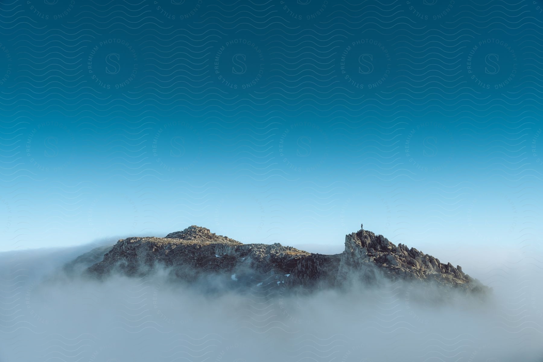 A person stands on the peak of a far-off mountain that is mostly surrounded by mist/fog beneath a blue sky.