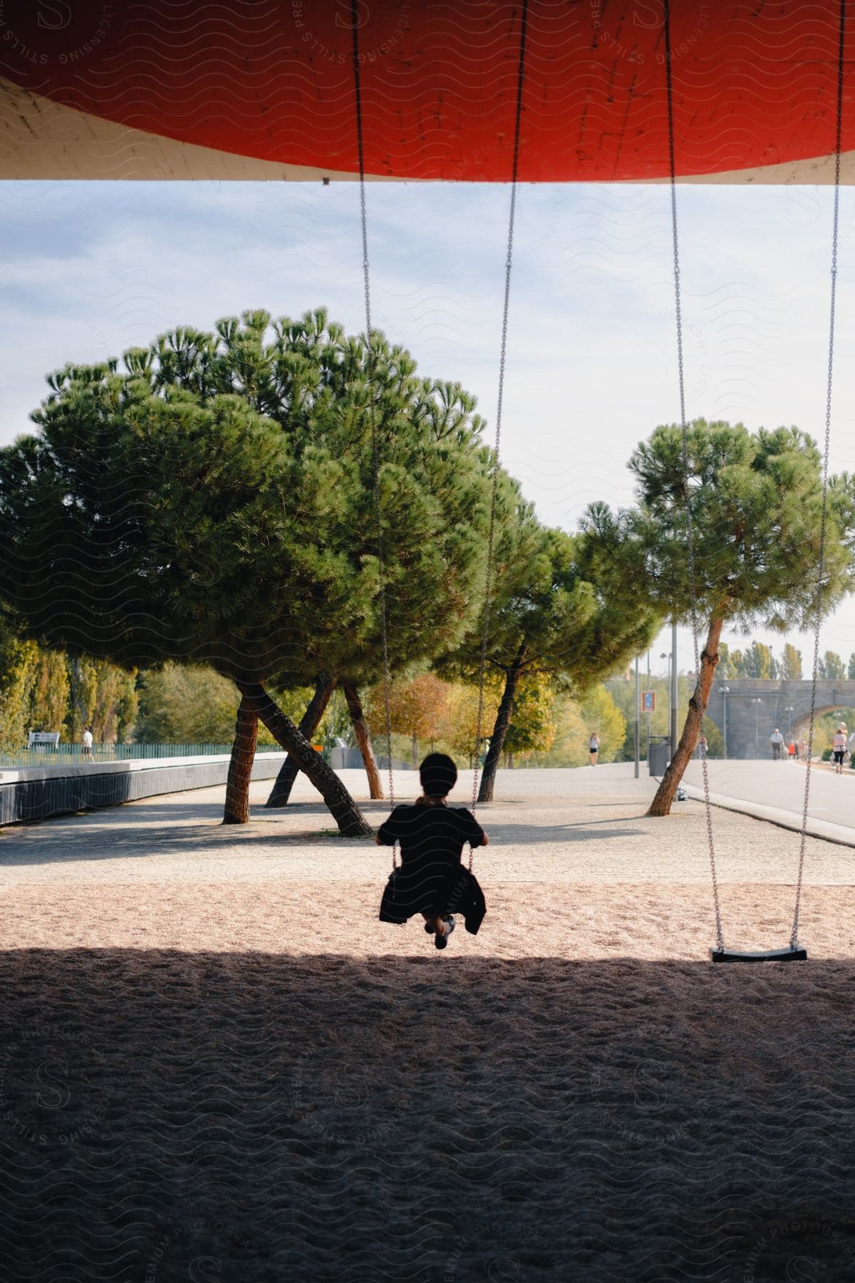 Silhouette of a woman on a swing in a public space with sand and trees.