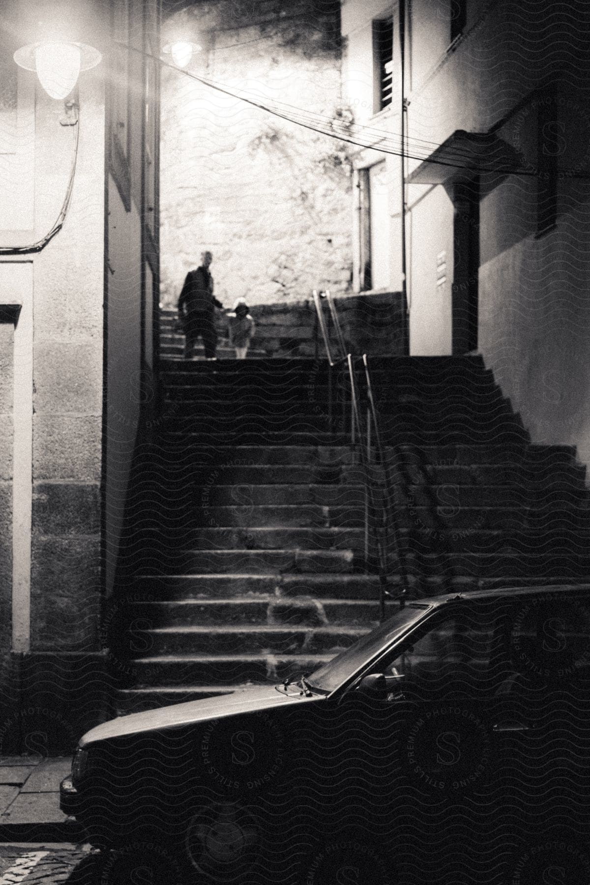 Black and white photo of a man walking down the stairs of a street with a child in a residential neighborhood and a car parked in front.