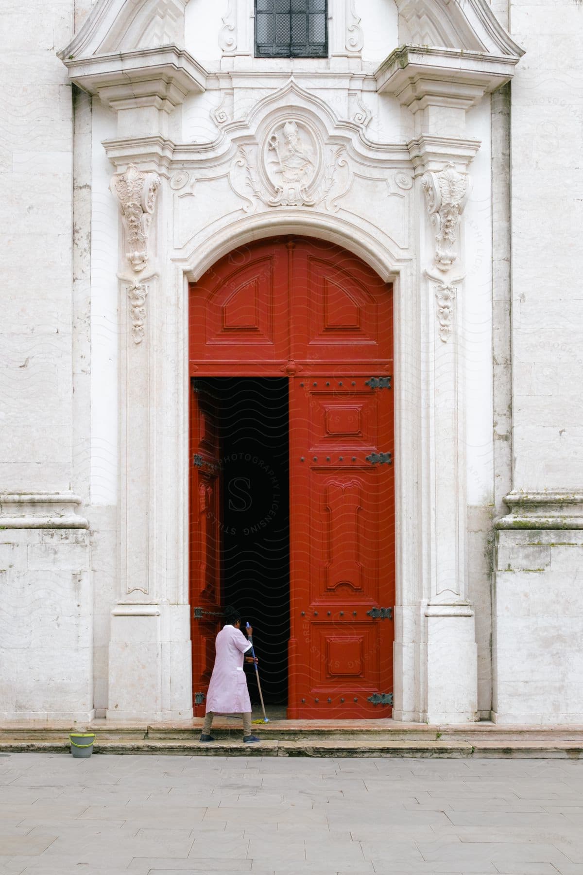 A woman diligently mopping the doorstep adorned with a big red door and surrounded by marble walls.