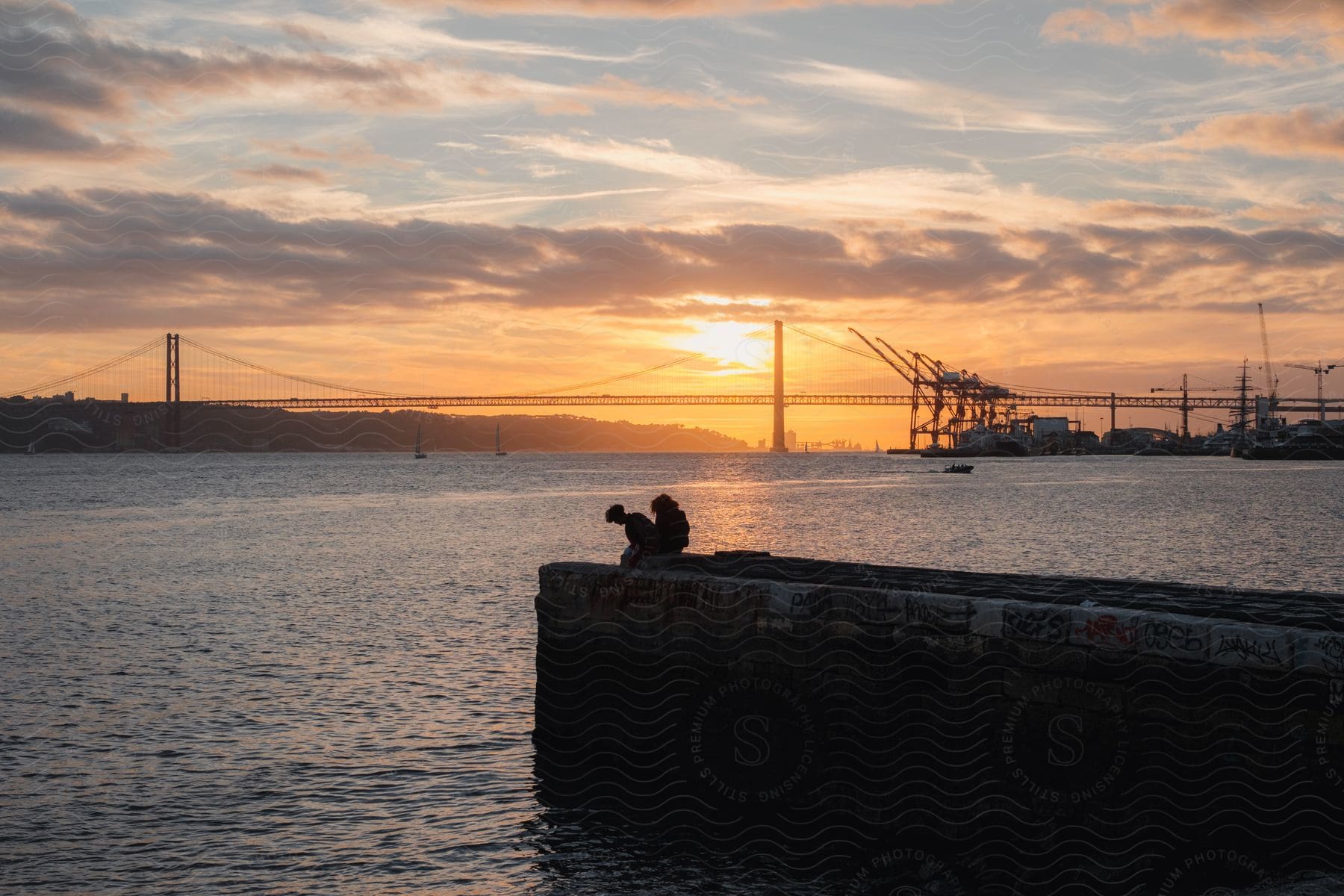 Two People Sitting On A Dock By The Sea With A View Of The Ponte 25 De Abril Bridge And The Sunrise On The Horizon