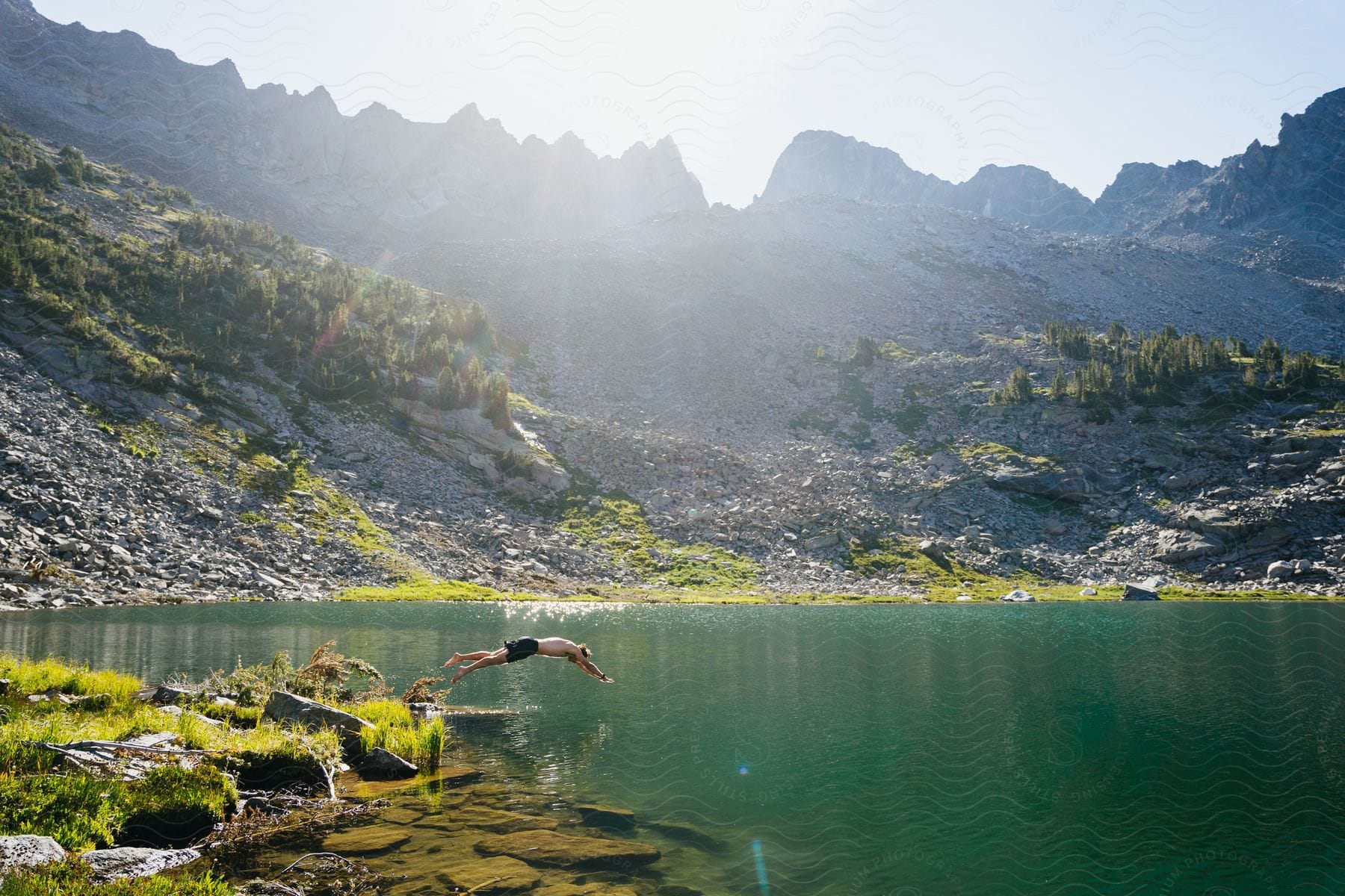 A man in shorts jumps into a serene lake, surrounded by a majestic mountain range.