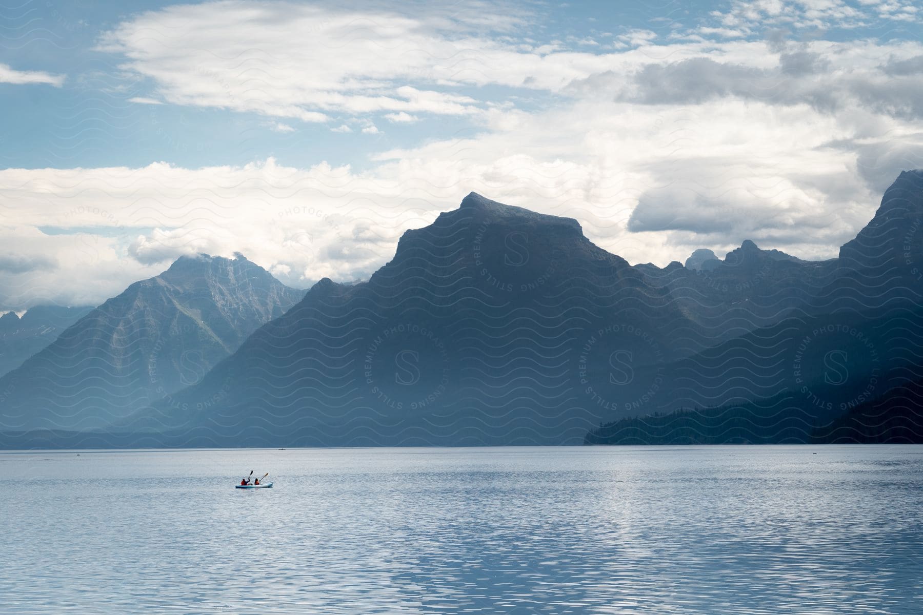 Panorama of the blue sea with people on the horizon in a kayak and a chain of mountains in the background.