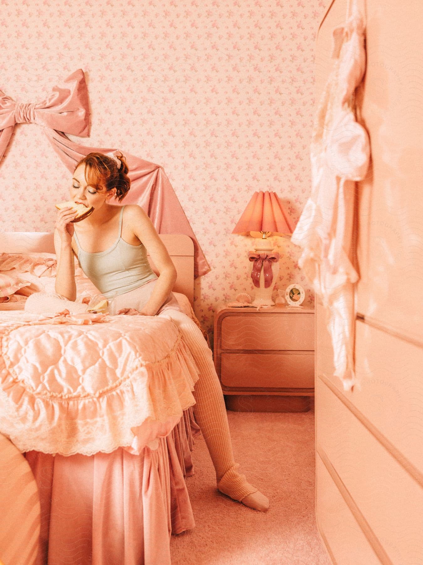 A woman in a ballet outfit sitting on a bed in a pink room, eating a slice of bread. The room is decorated with pink wallpaper, a large bow, and vintage furnishings.