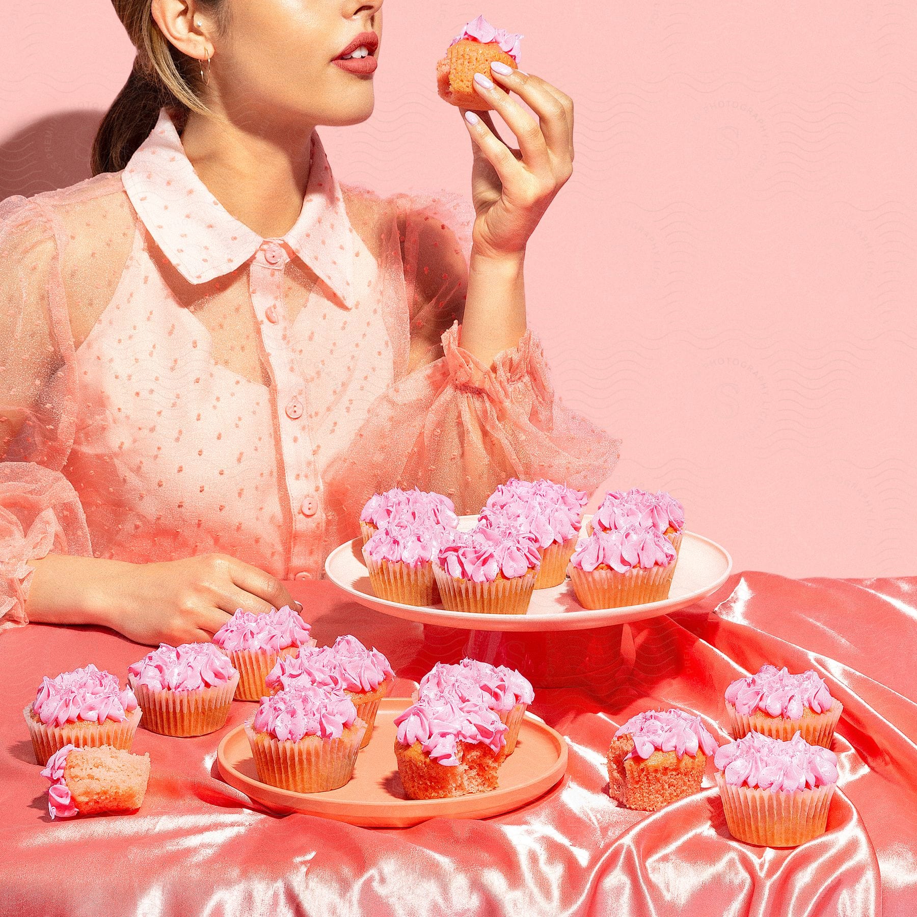 Stock photo of a woman in a pink outfit with a translucent puffed sleeve, holding and eating a cupcake, with plates of pink frosted cupcakes on a pink satin tablecloth.