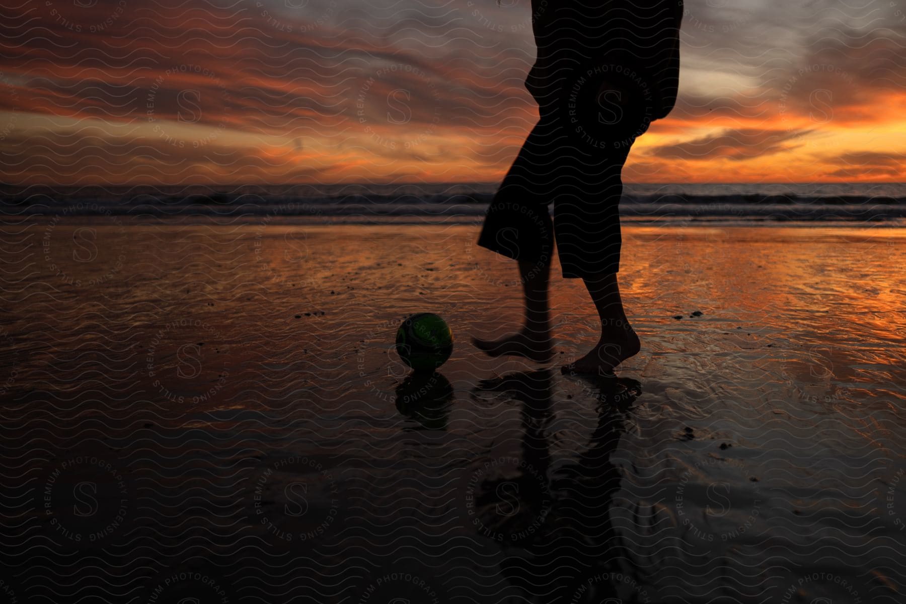 View of a person's legs with a ball in the sand of a beach in an orange dawn sky.