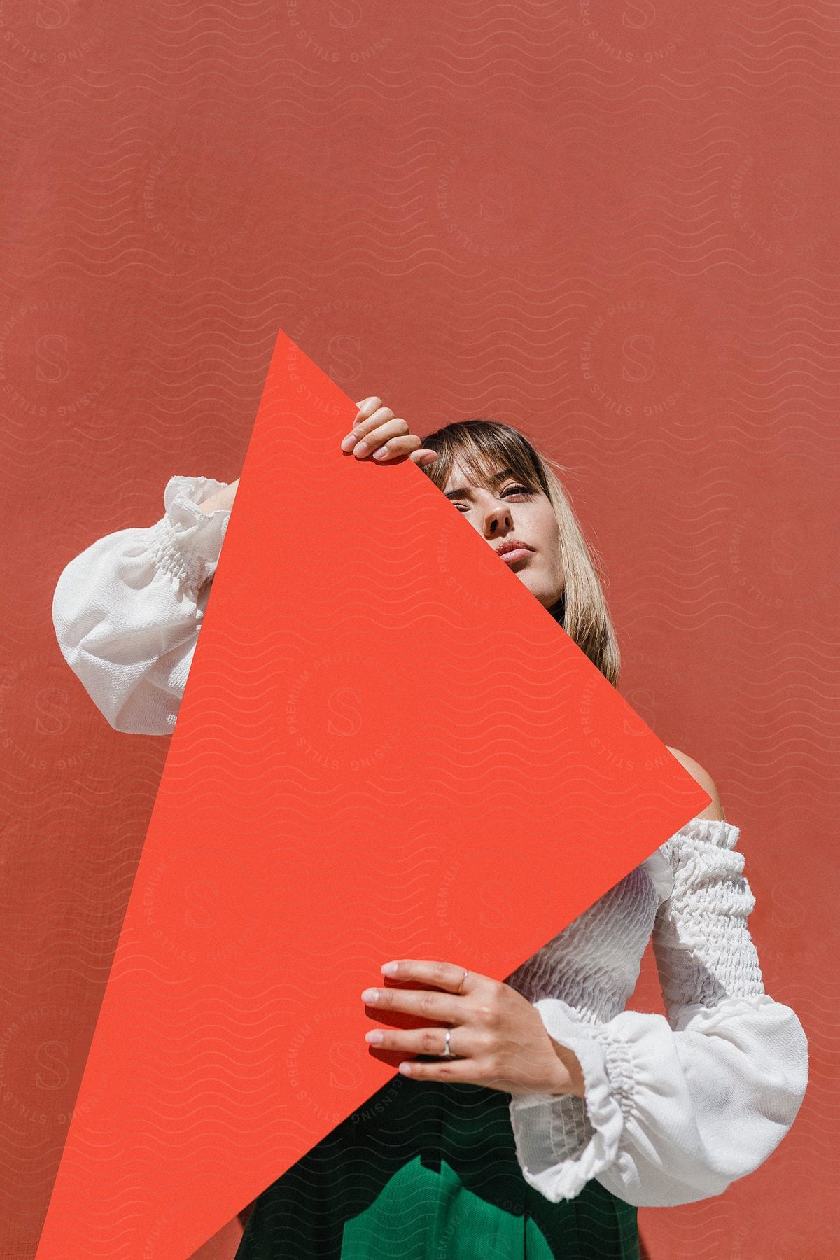 A blonde woman, dressed in a white blouse and green pants, holds an orange triangle paper against a backdrop of a matching orange background