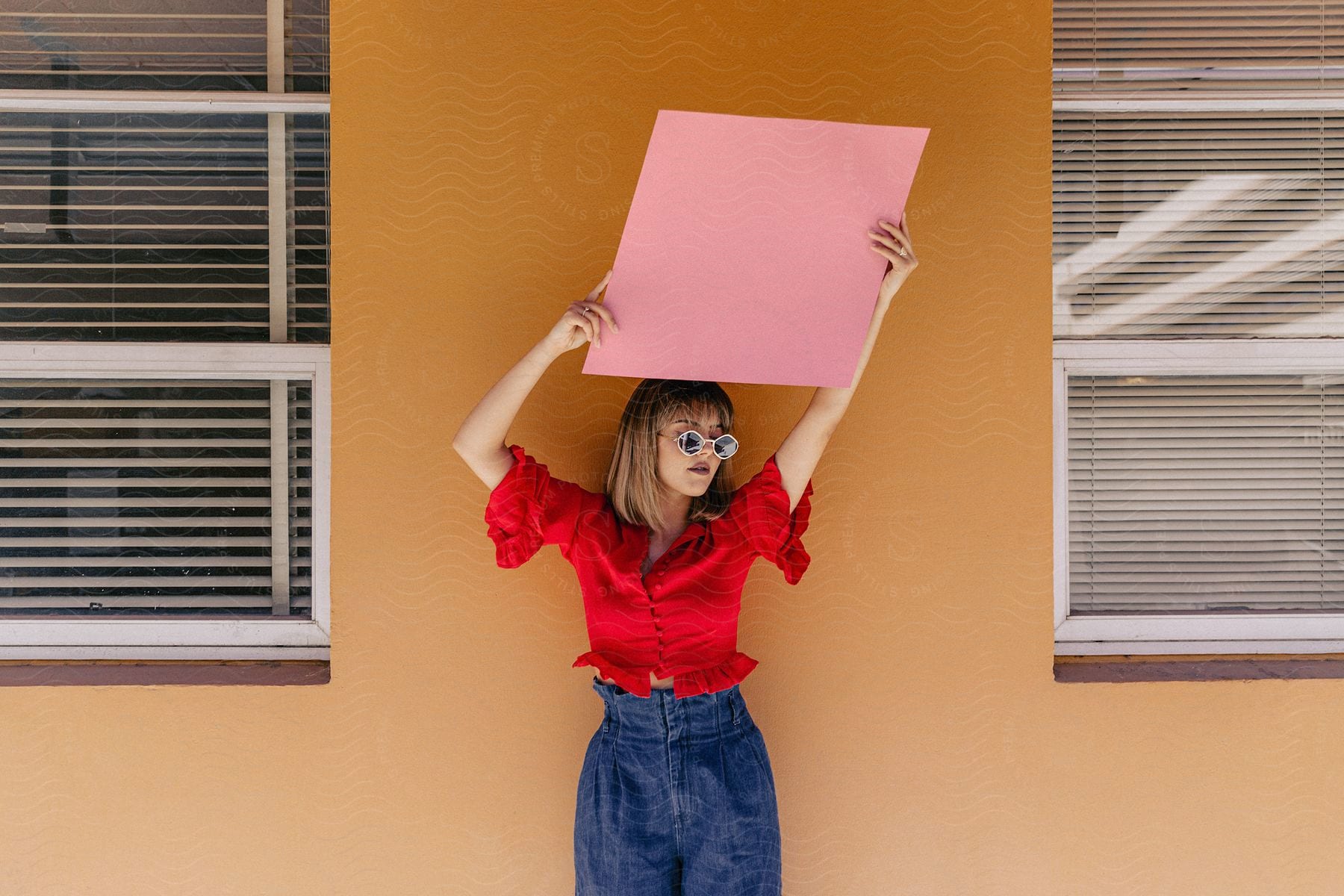 A woman with brown hair, sunglasses, a red blouse, and jeans holds up a pink paper in front of an orange wall between windows