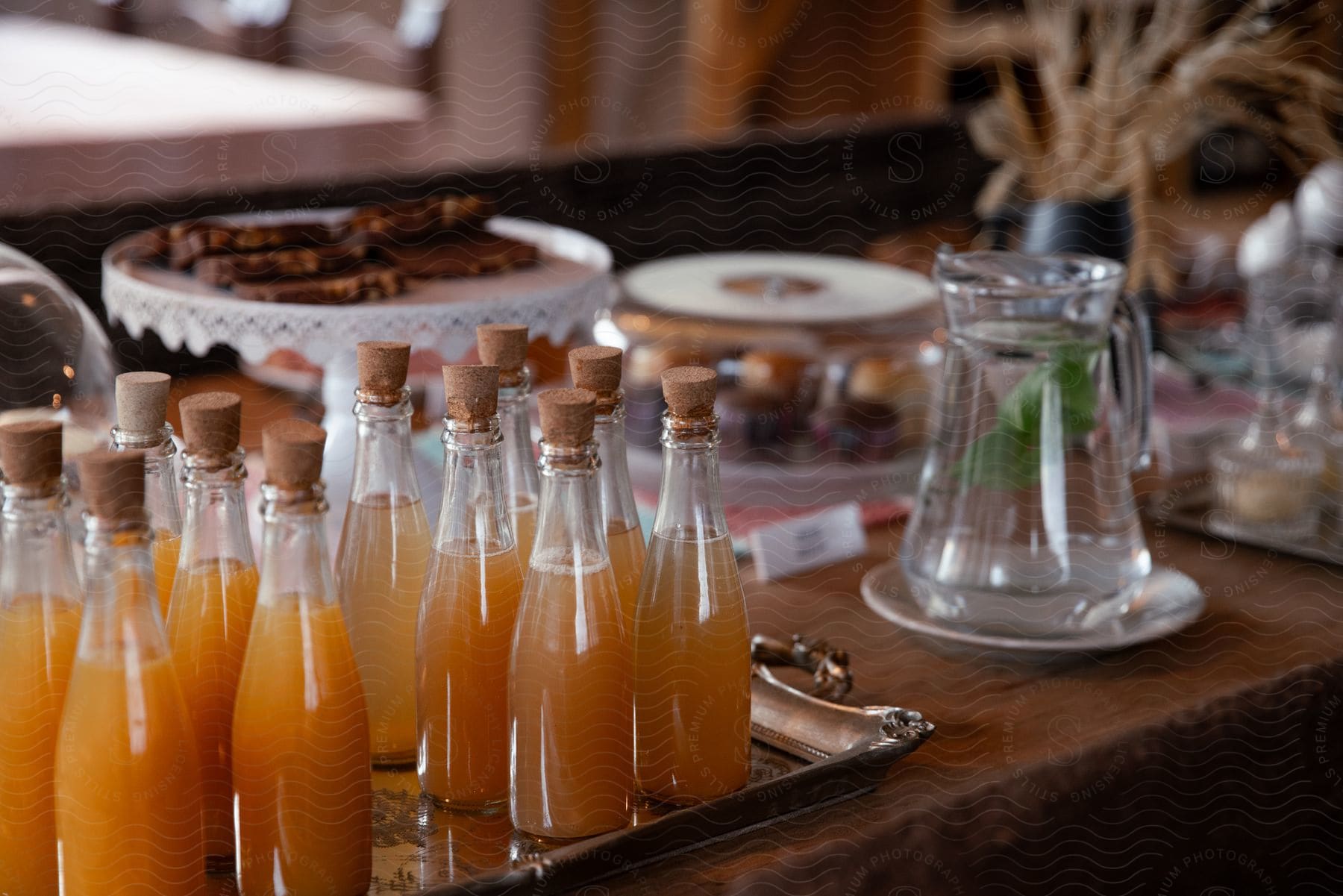 Rows of bottled orange beverages on a tray, with dessert trays and a water jug with fresh mint inside in the background, on a wooden table.