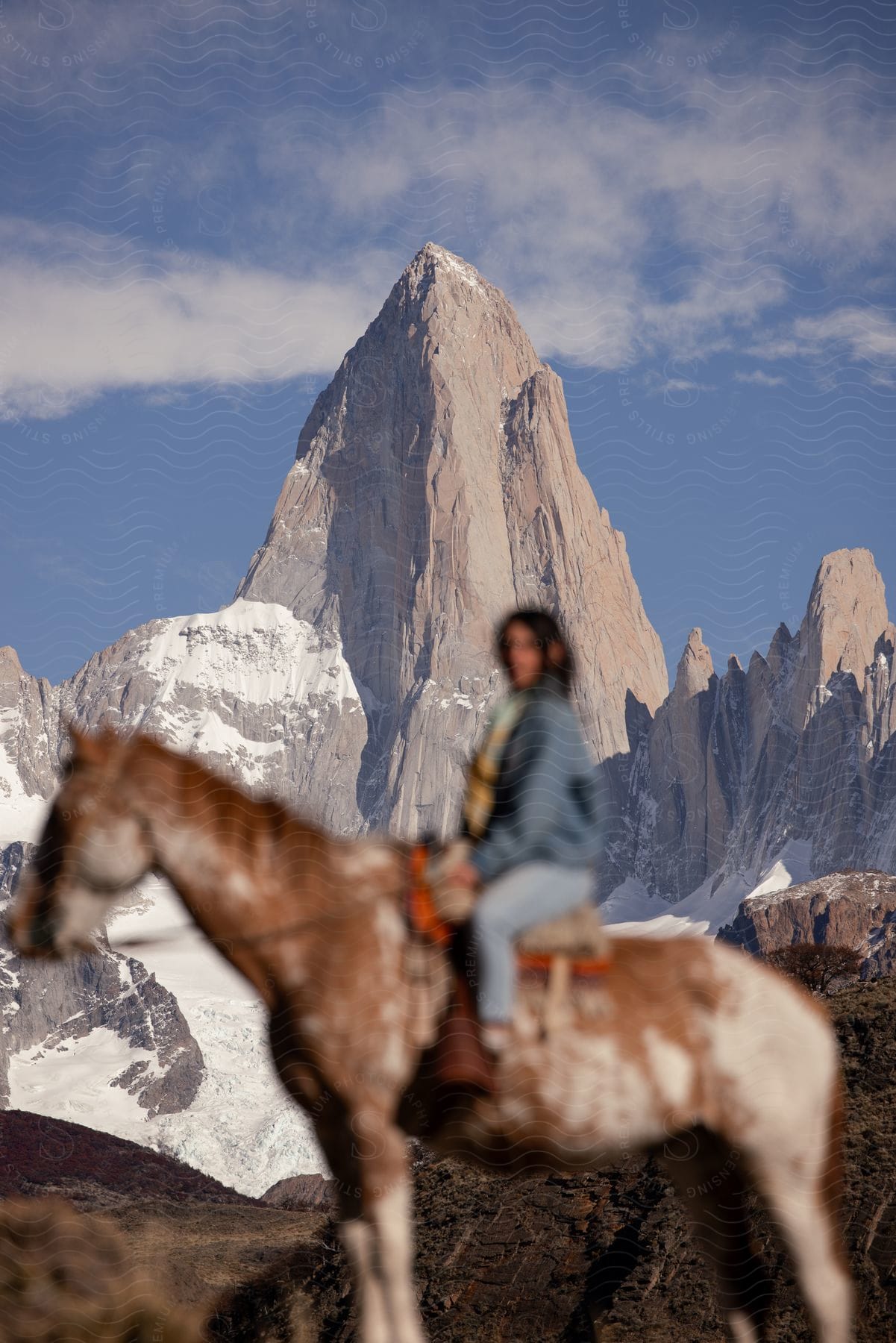 Woman on horseback out of focus, with a sharp mountain peak in the background under a blue sky.