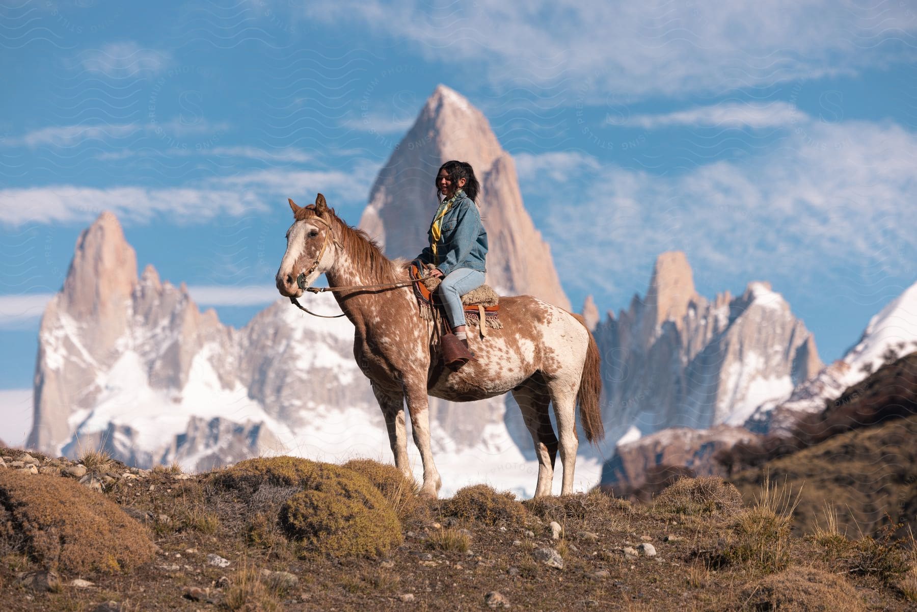 A woman gracefully rides her horse atop a majestic snow-capped mountain, framed by a captivating sky adorned with billowing clouds.