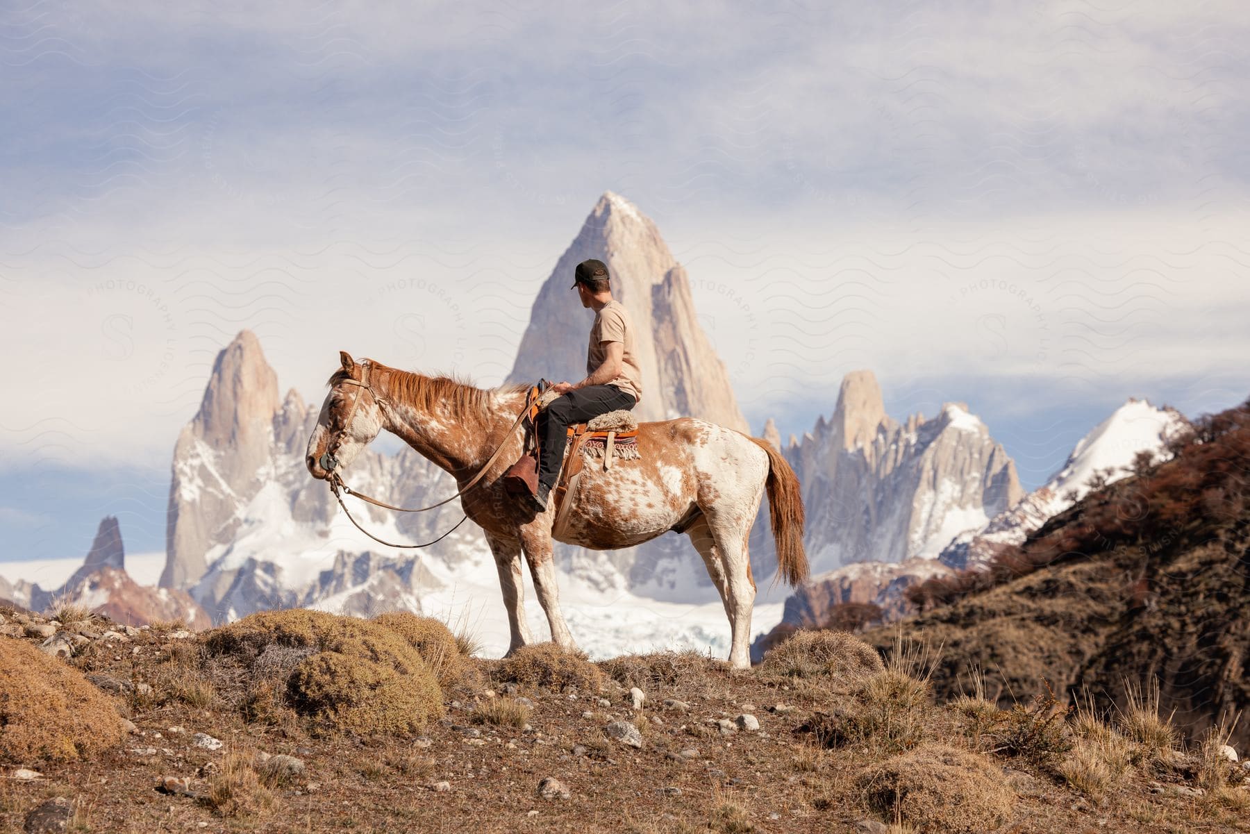 A man sits on top of a horse and stares at the steep mountain range behind him.