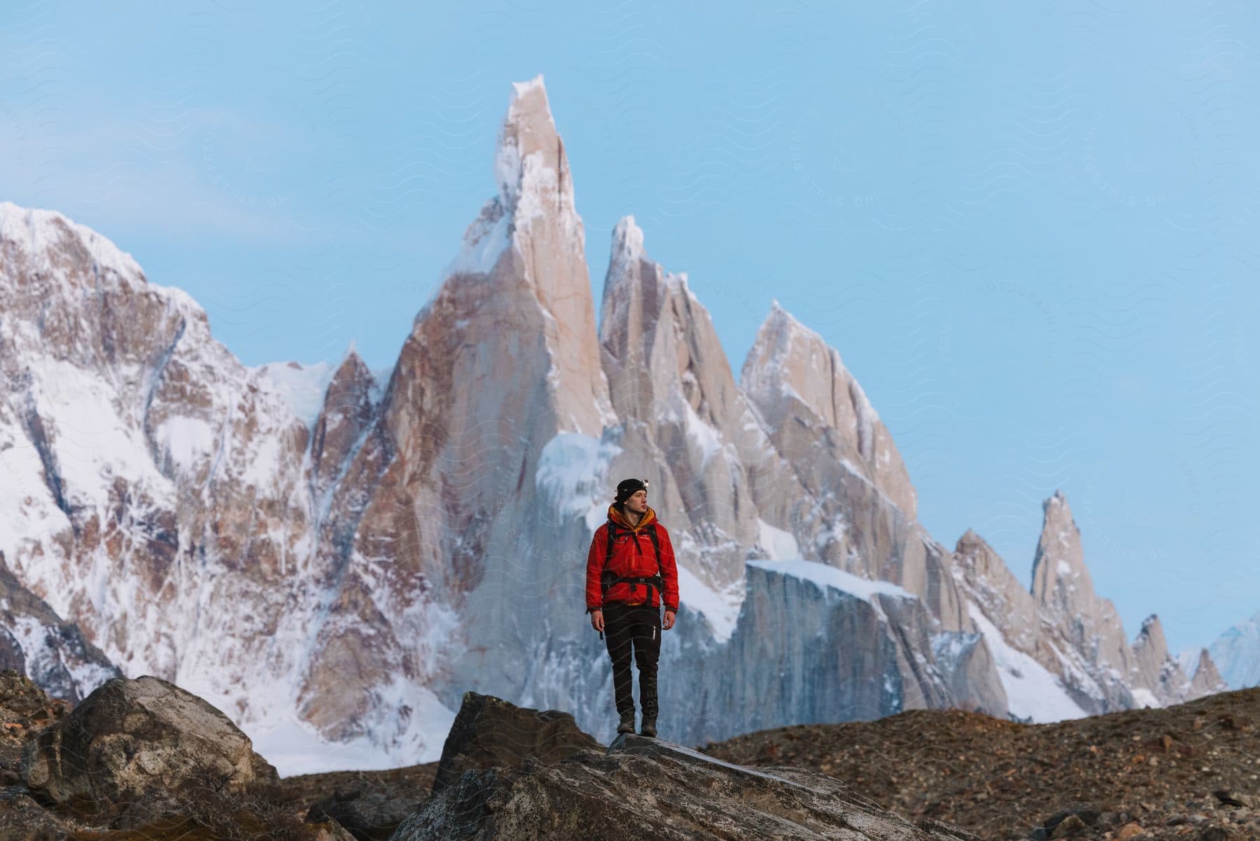 A rugged adventurer, clad in a crimson jacket and carrying a backpack, stands atop a towering rock, gazing upon a majestic snow-covered mountain range under a vast, pristine sky