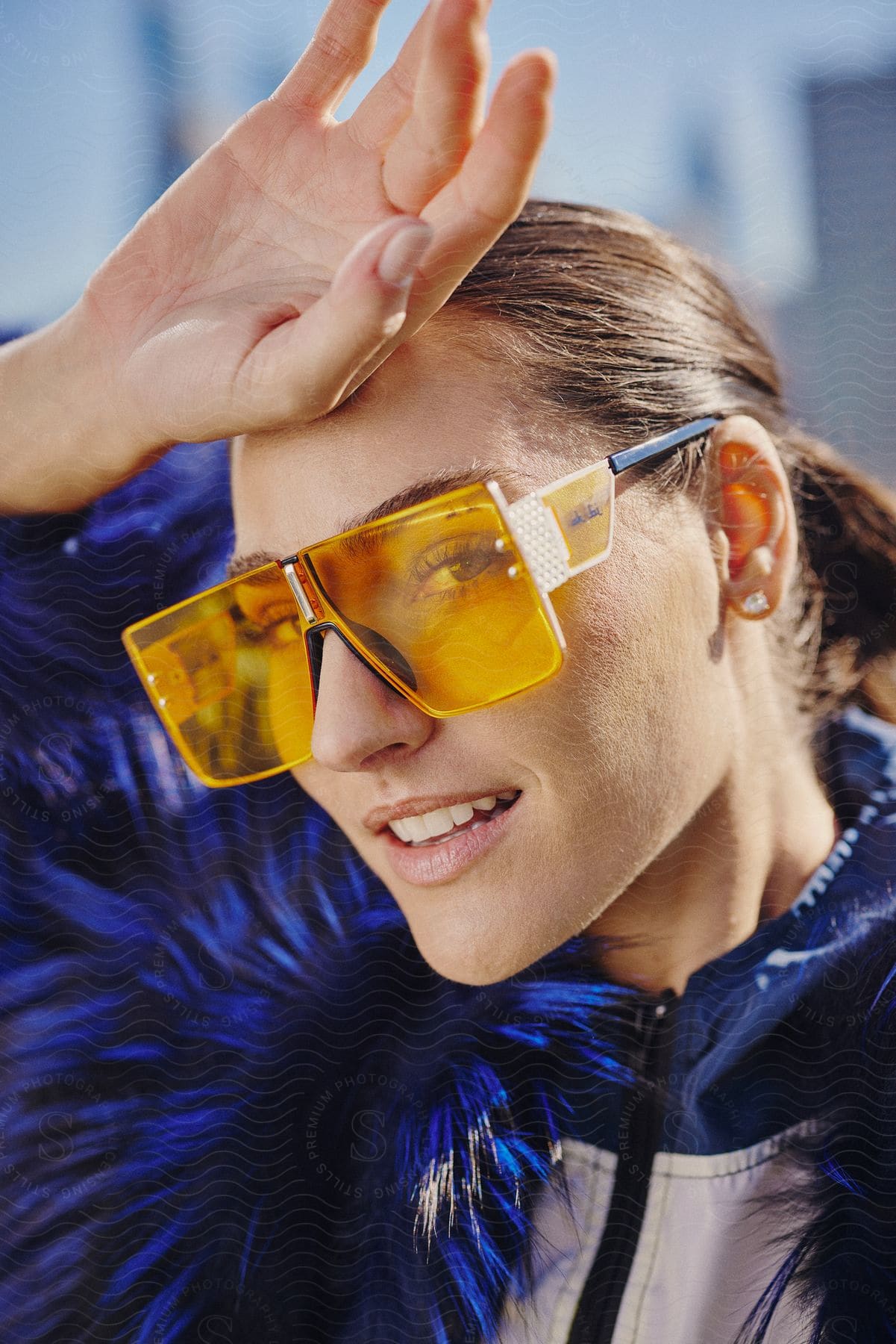 A young woman wearing protective glasses and a blue jacket smiles as she holds her hand over her forehead