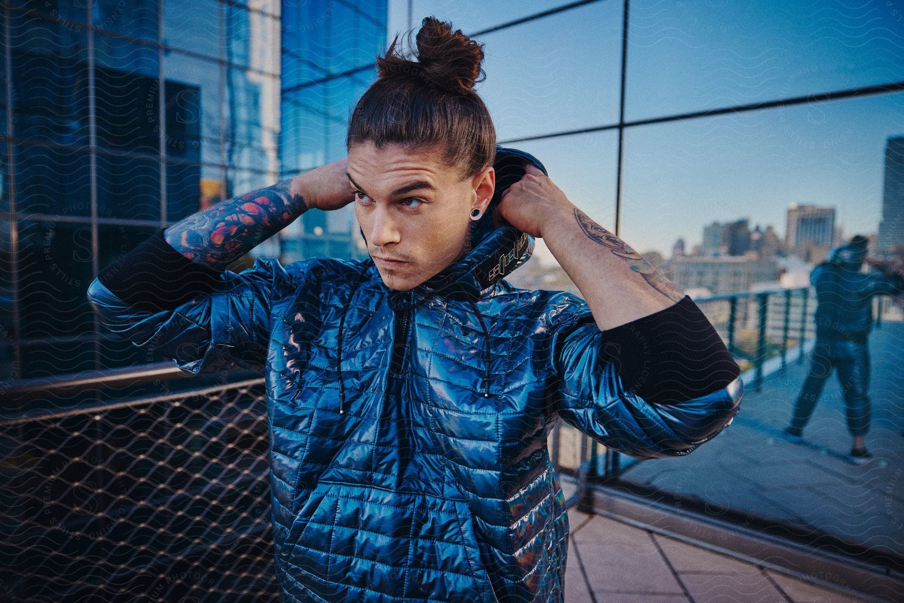 Person with tattoos adjusting the hood of a blue jacket in an urban setting with reflective glass buildings.