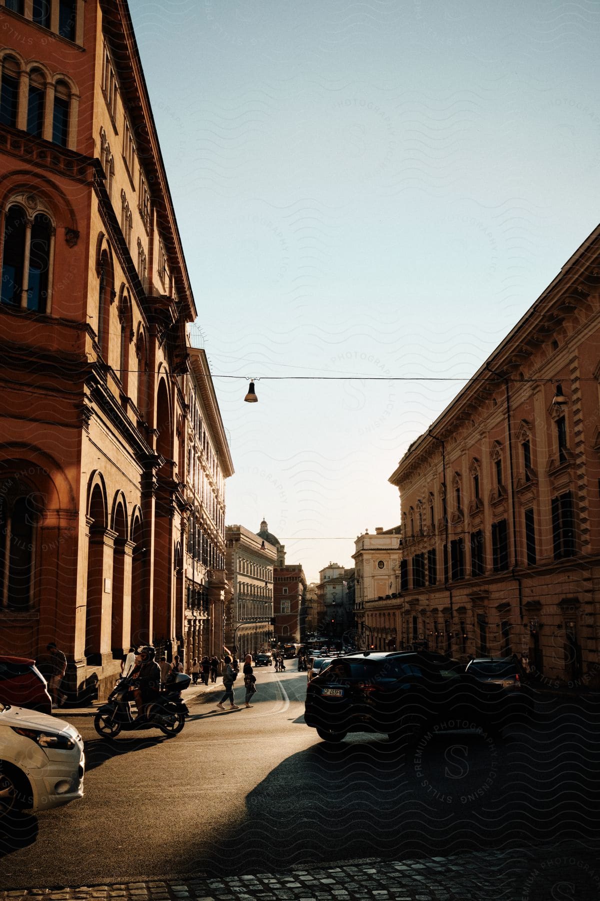 Pedestrians walk along street between architecturally detailed buildings near a busy intersection.