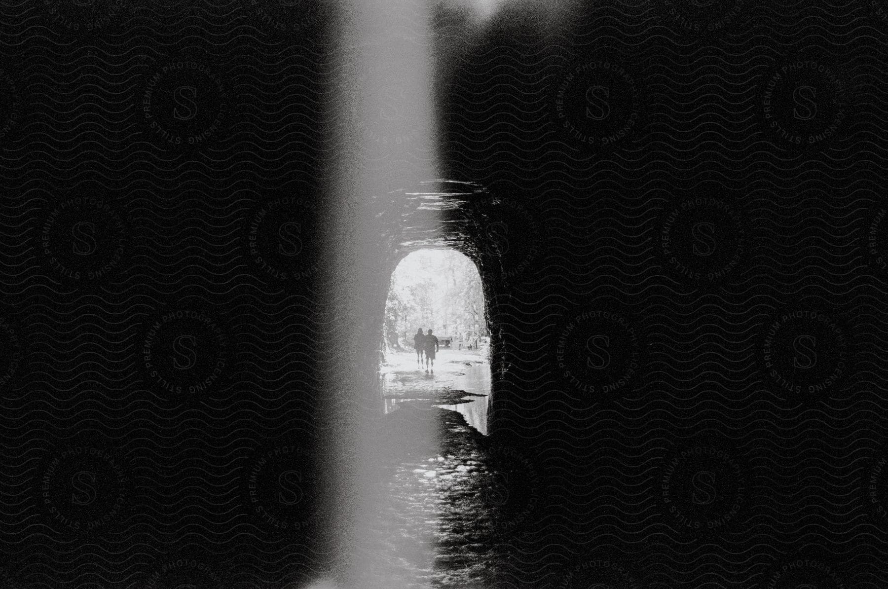 View from inside a cave with the silhouette of two people on the outside walking under the flowing water of a river.