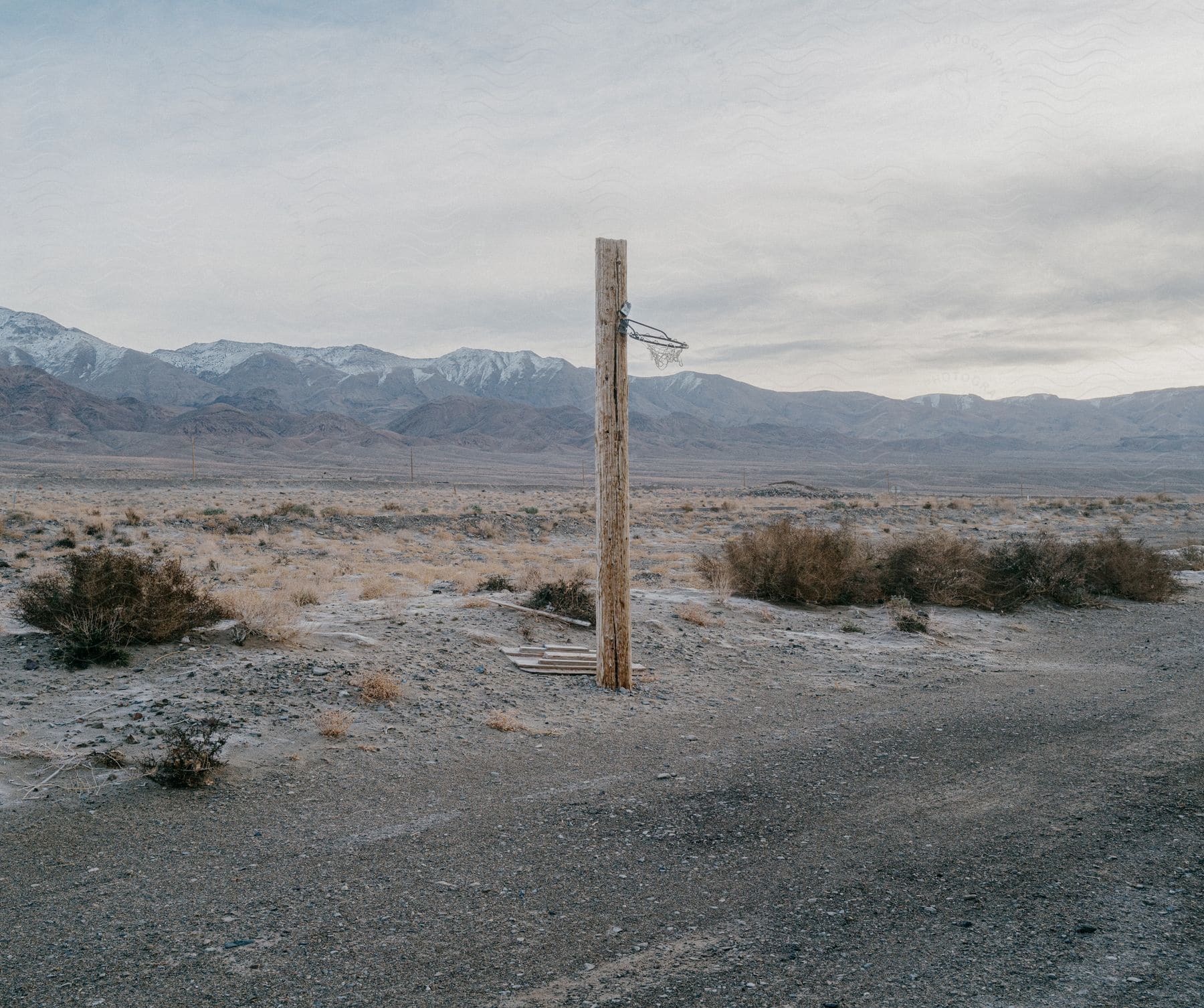 A weathered basketball hoop atop a lone wooden post stands out against the backdrop of a vast plain and distant mountains under a gloomy sky.