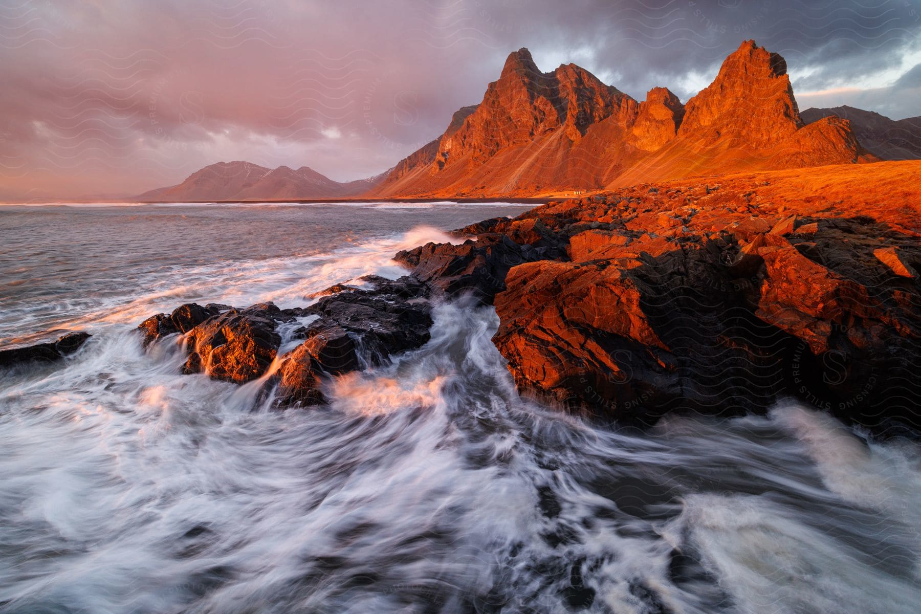 The rocky shore explodes with white-capped waves crashing against the cliffs, set against the fiery orange backdrop of the setting sun and majestic mountains in the distance.