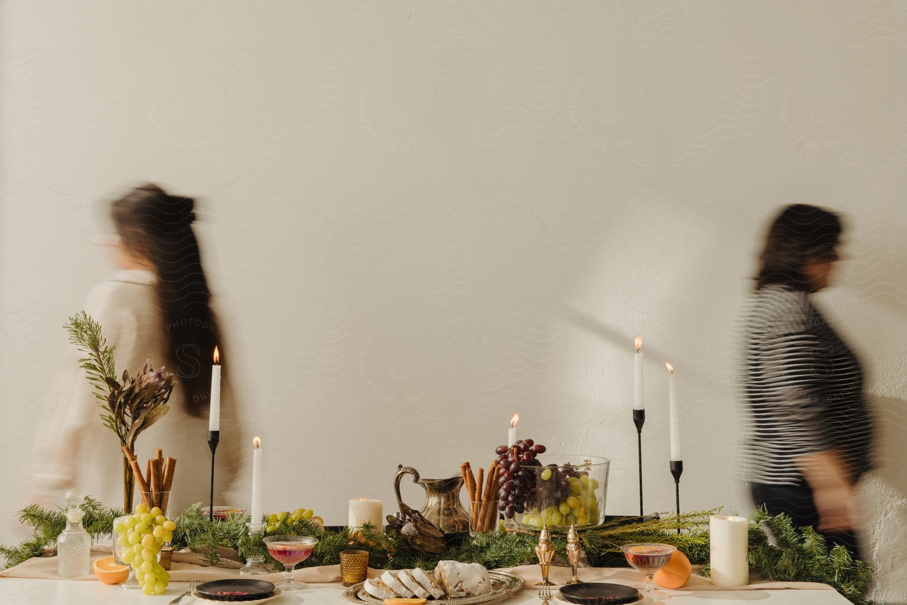 Two women pass a candlelit table laden with food, one approaching, the other leaving.