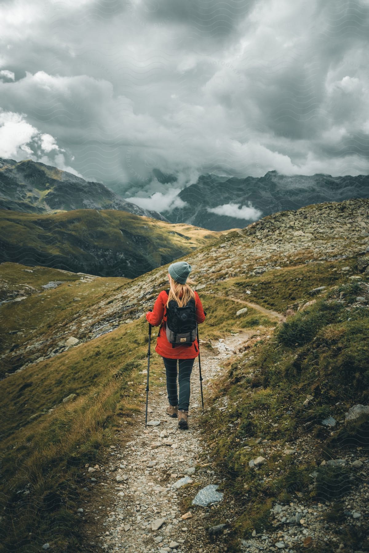 A blonde woman, donning a gray hat, an orange jacket, a black backpack, and holding trekking poles, walks down a dirt track in a field with mountains under clouds in the background.