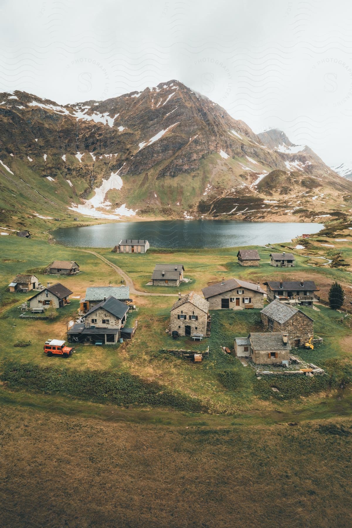 Aerial panorama of farmhouses in a grassland facing a lake amidst a chain of snow-covered mountains on a cloudy day.