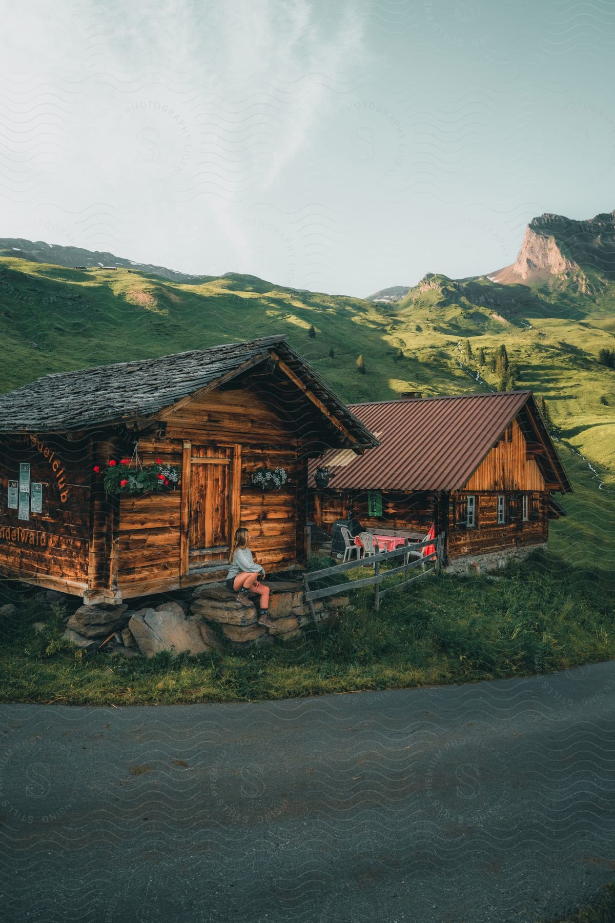 Woman sitting at the entrance of a cabin in a natural setting with hills and mountains.