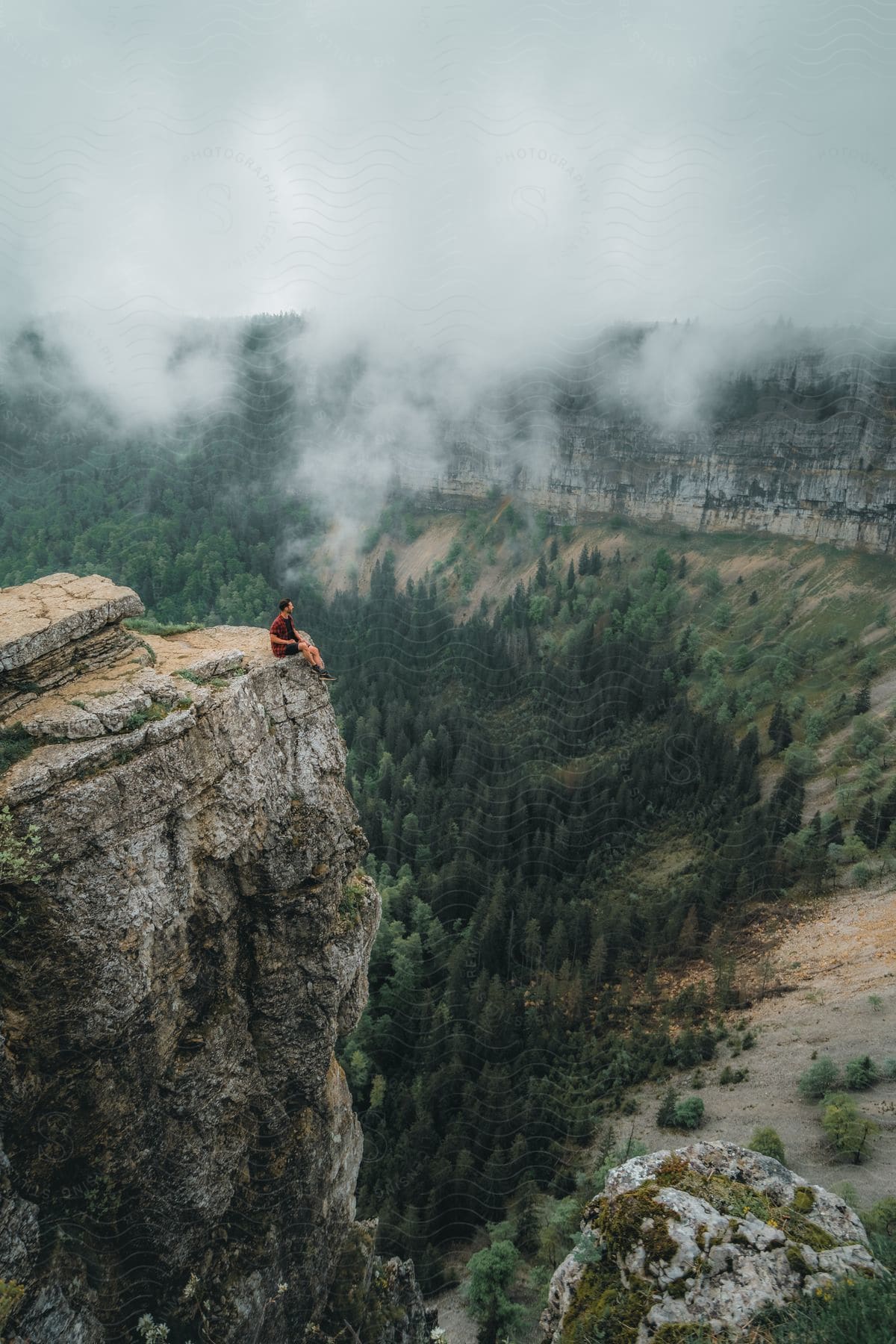 a person sitting on a cliff overlooking a valley and forest in the foggy weather
