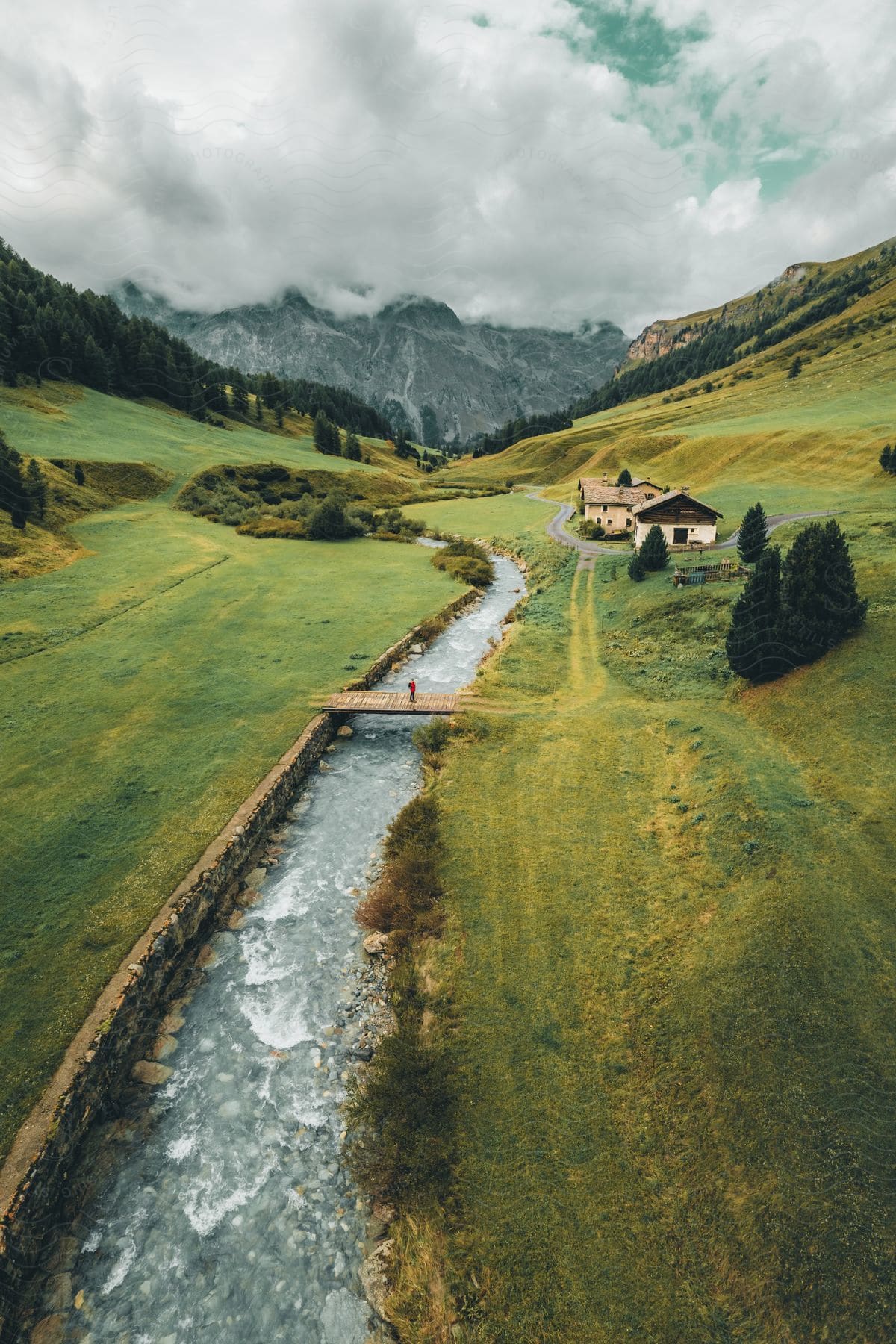 Stock photo of an aerial view of a person crossing a wooden bridge over a river, with houses lining the bank in a picturesque green valley.