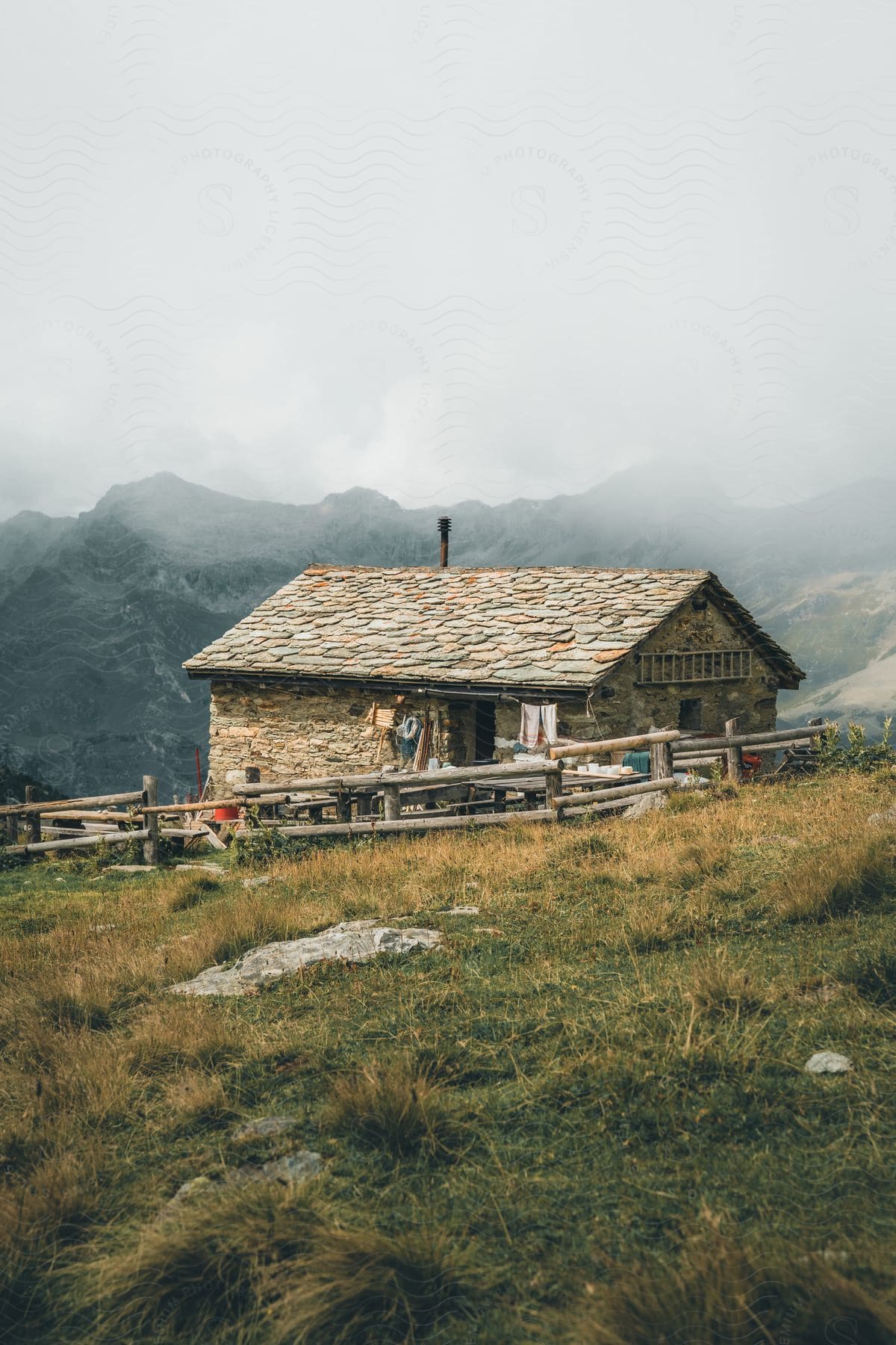 A cozy stone cabin nestled on a mountain hill, surrounded by a rustic fence and shrouded in misty peaks.