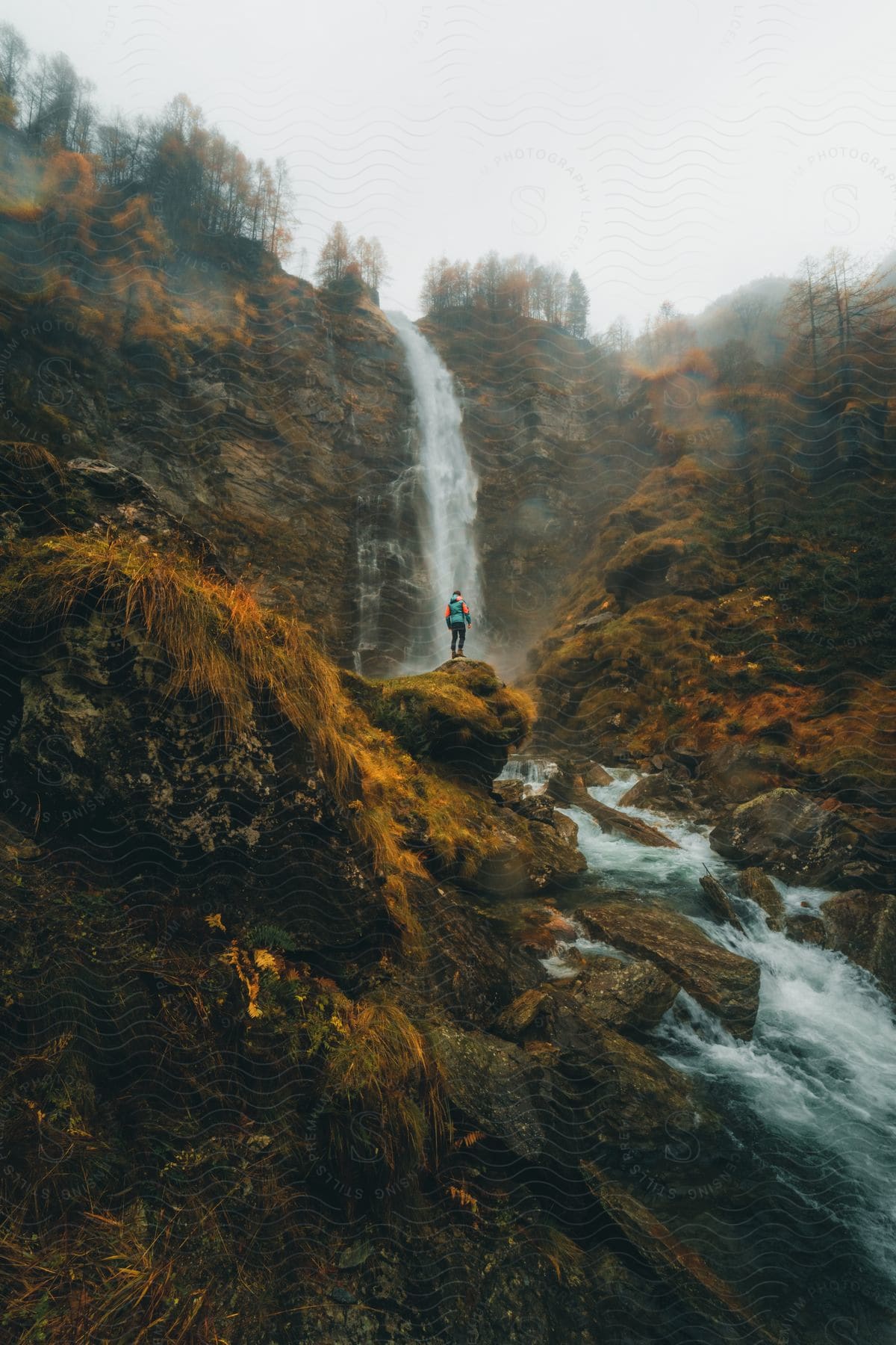 Landscape of a person on a cliff facing a waterfall on a cloudy day.