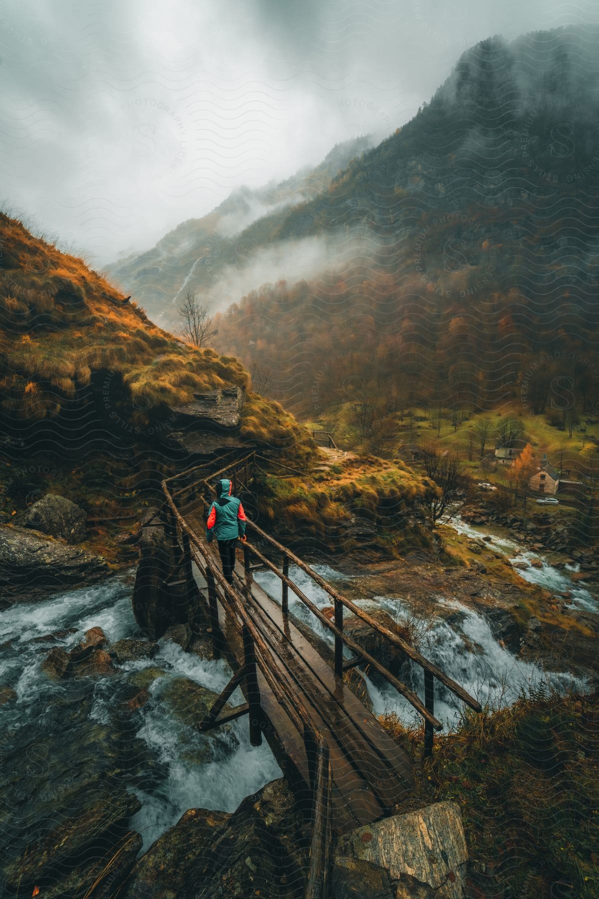 A lone figure on a mountain bridge watches the river dance beneath, autumn's fiery breath painting the distant peaks with vibrant hues, all shrouded in a misty embrace.