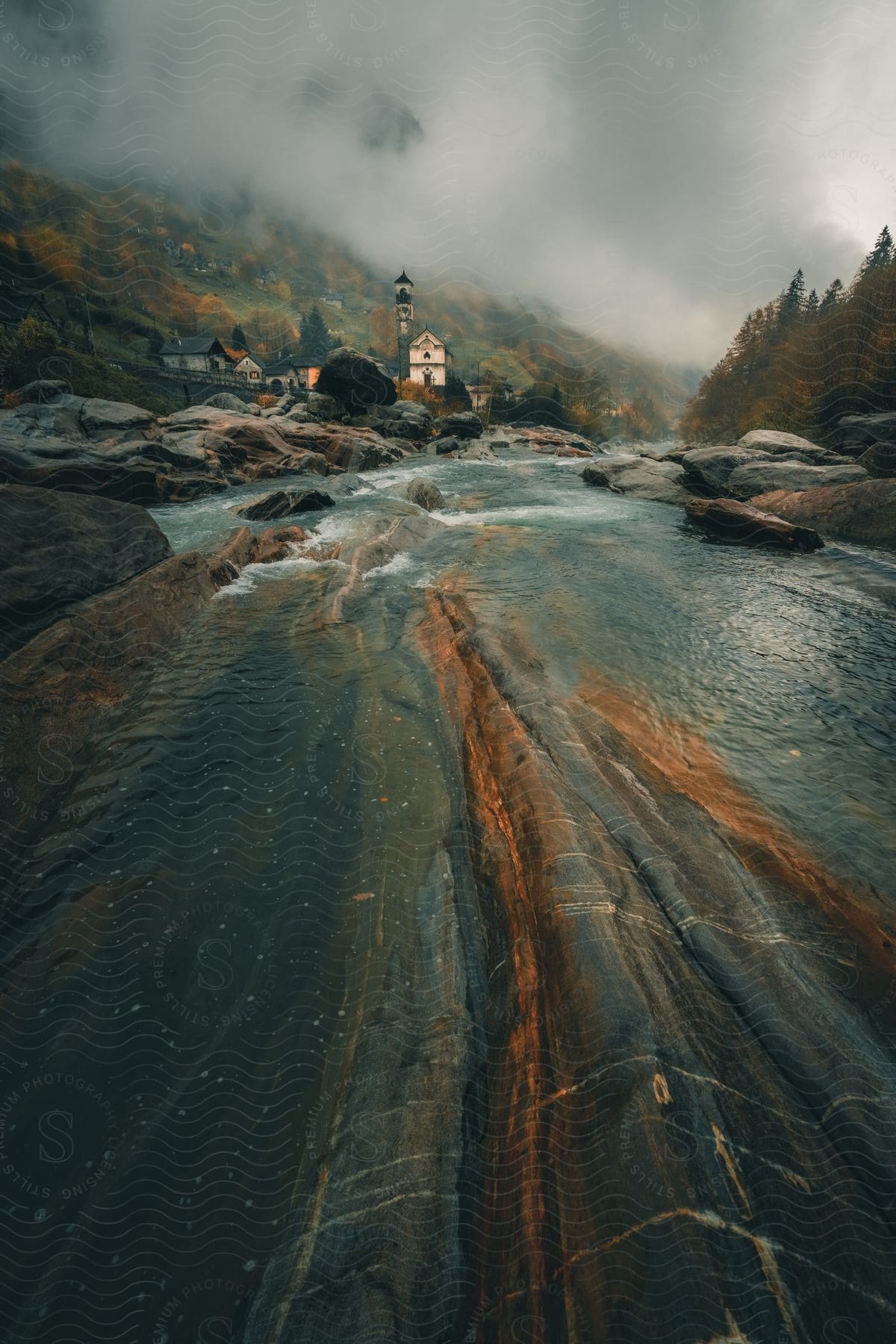 Mountain stream flowing over rocks with a church in a misty village background.