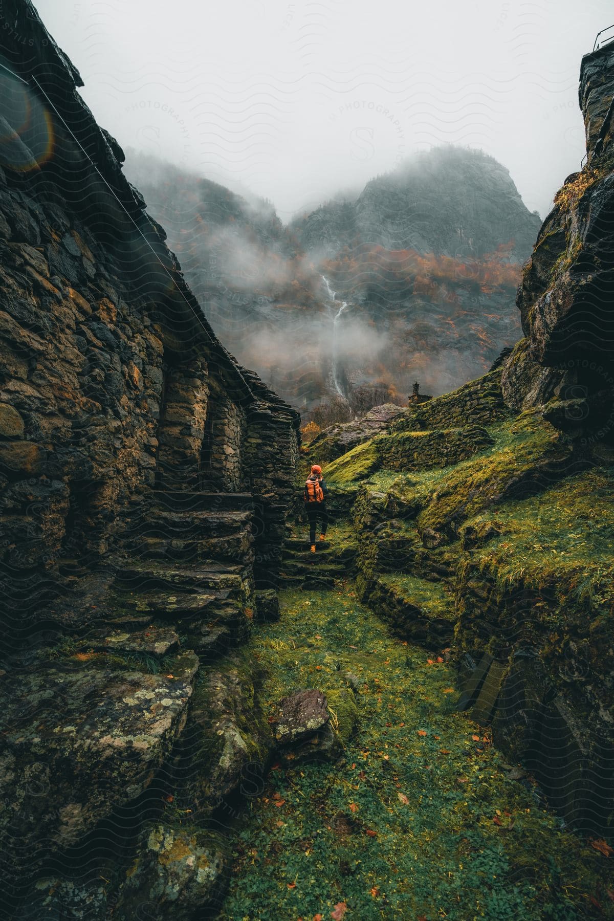 A hiker in a bright orange jacket surveys a misty mountain vista from atop a grassy knoll, a weathered stone structure standing sentinel beside them.