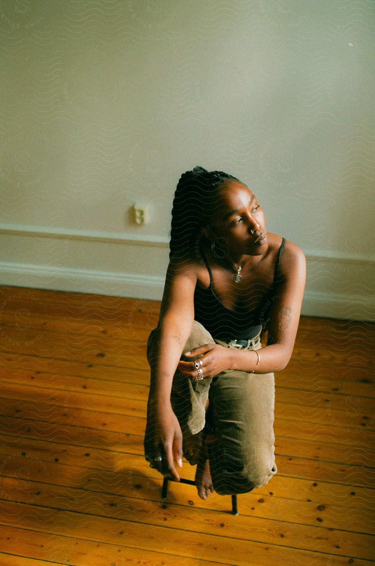 A woman sitting on a wooden stool in a room with hardwood floors, looking to the side.