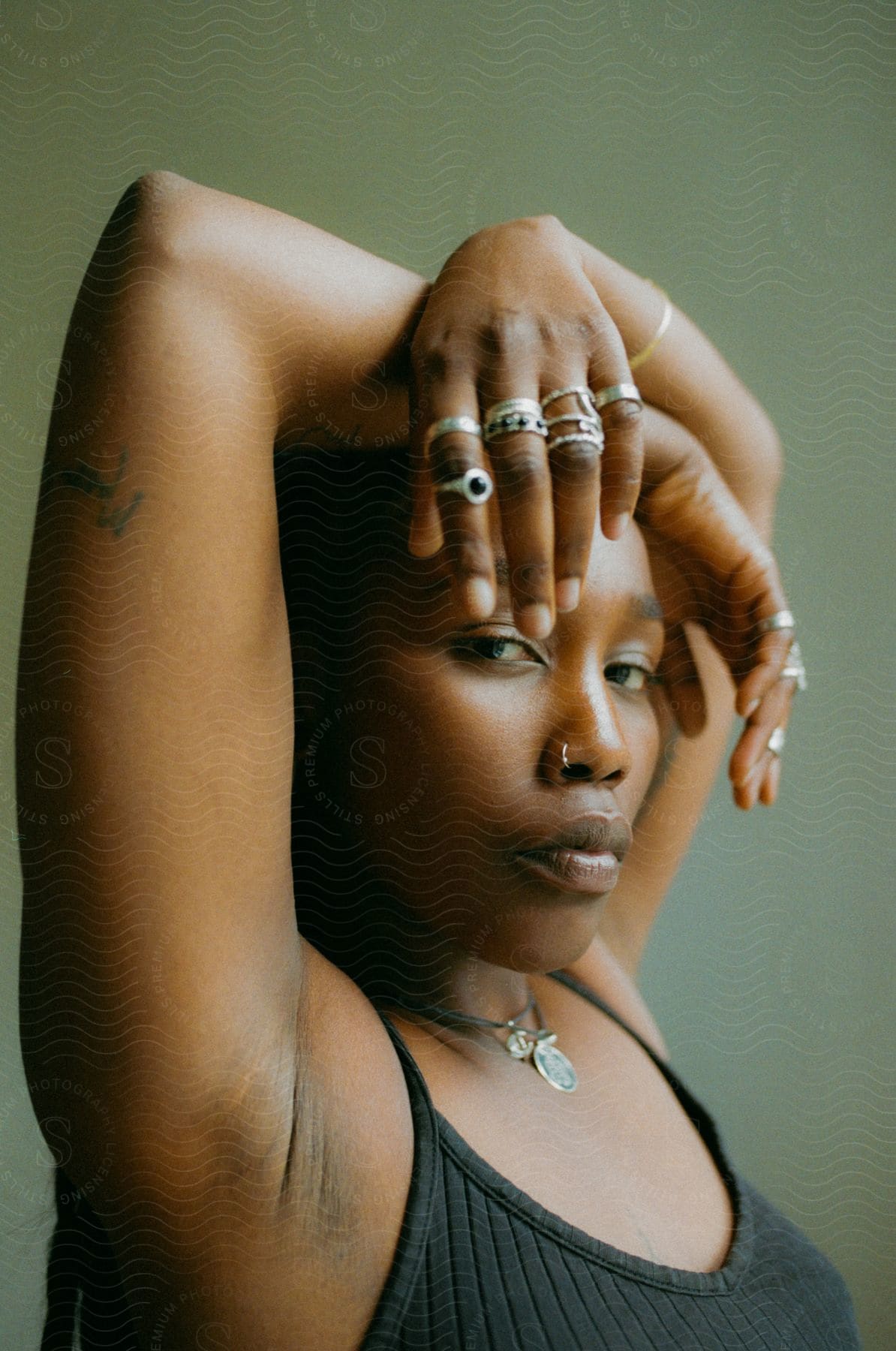 A woman, donning a black shirt, necklace, nose piercing, and rings on her fingers, confidently poses for the camera.