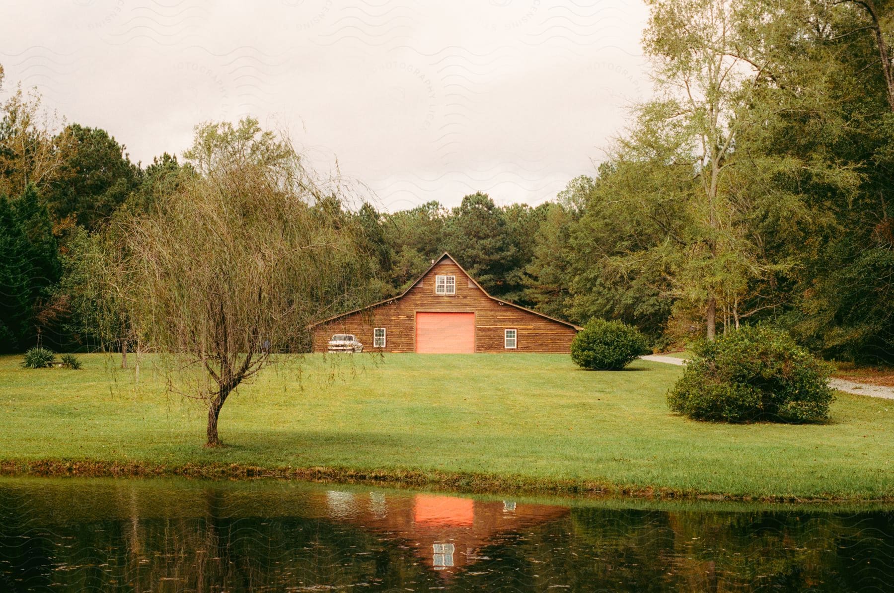 A wood plank farm house with a long driveway and lawn near a pond