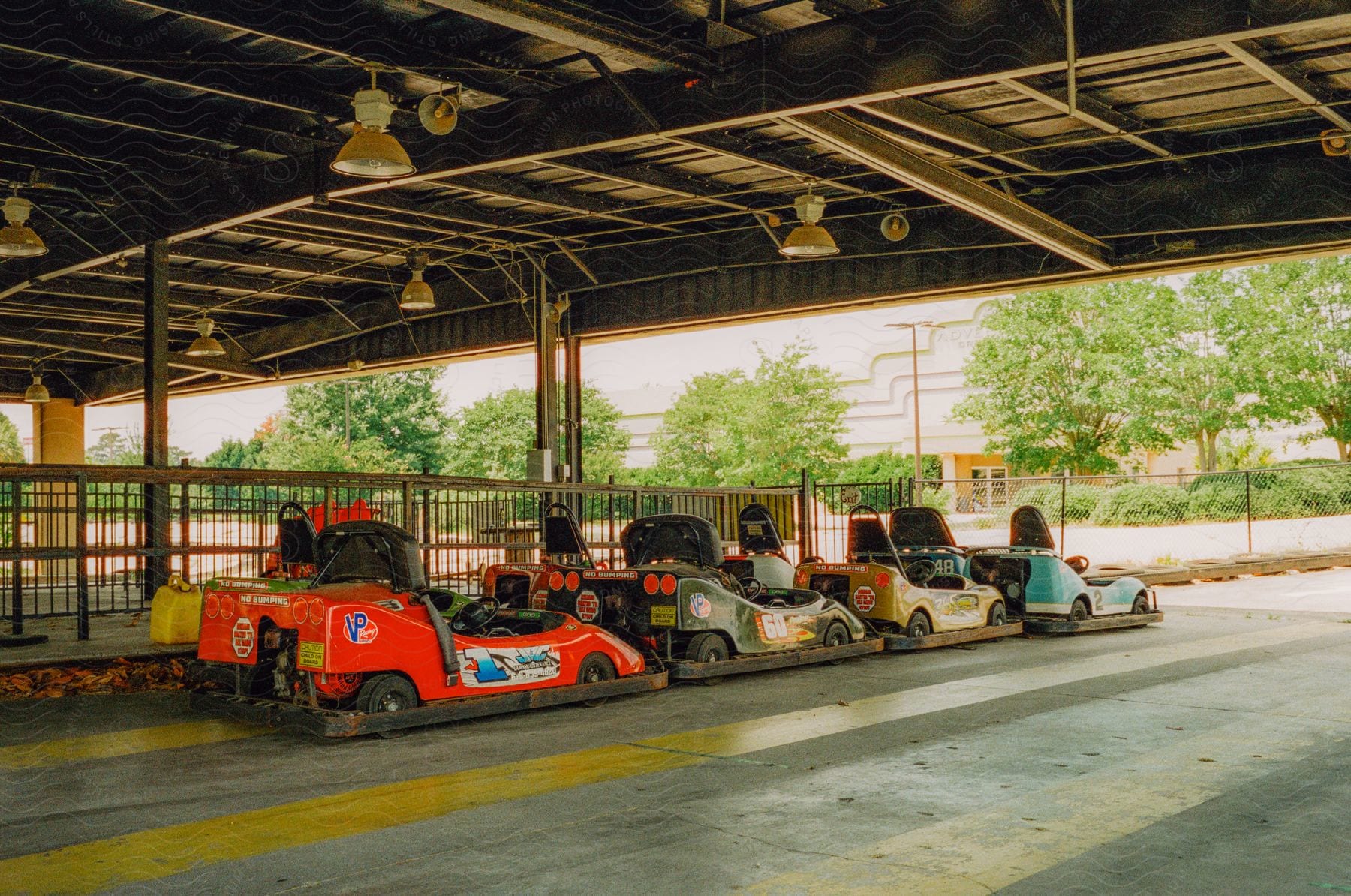 Bumper cars are lined up along a fence with trees and a building in the distance