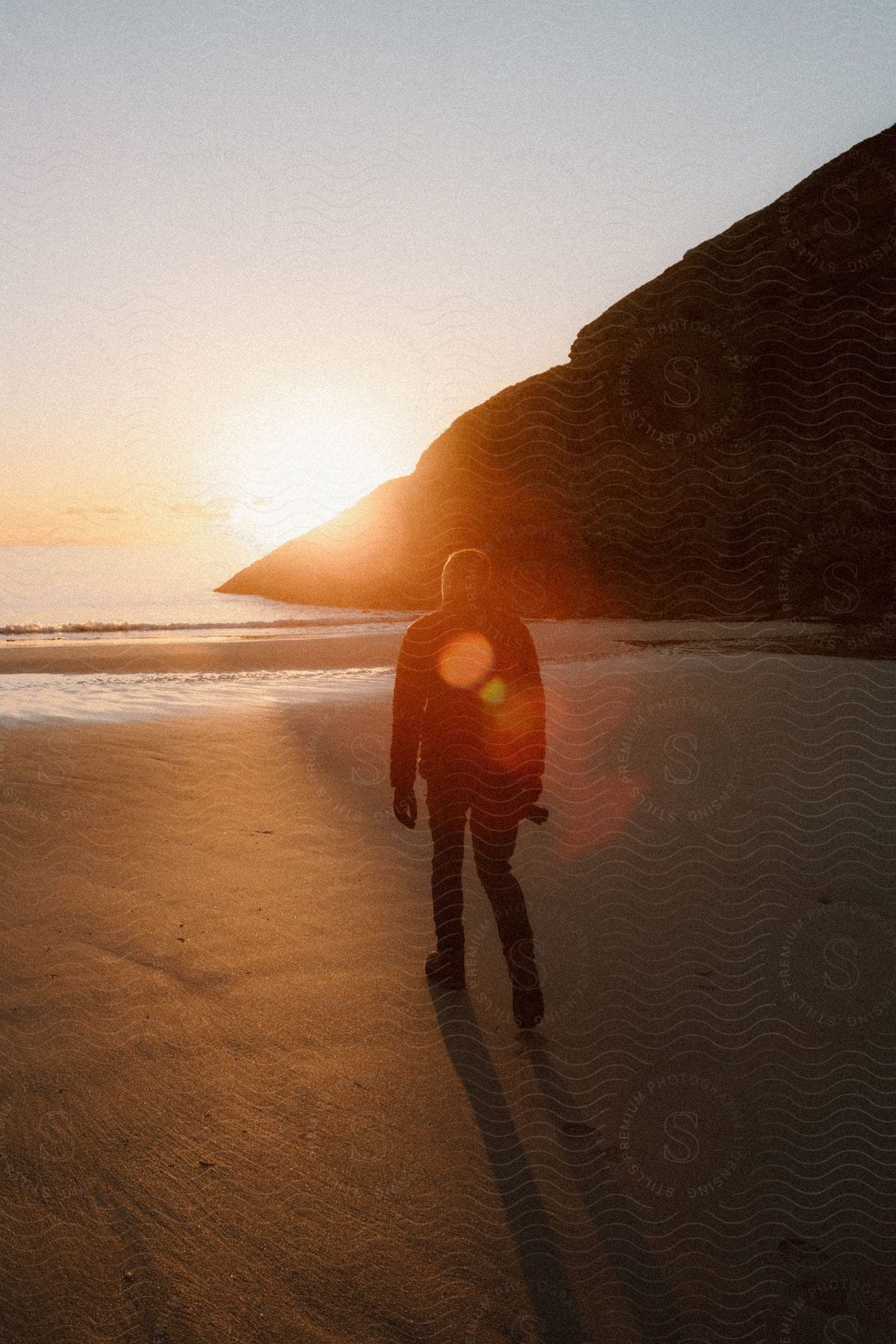 A man, dressed in a black jacket, pants, gloves, and boots, carries a camera on the beach as the sun sets, capturing the scenic beauty of the moment.