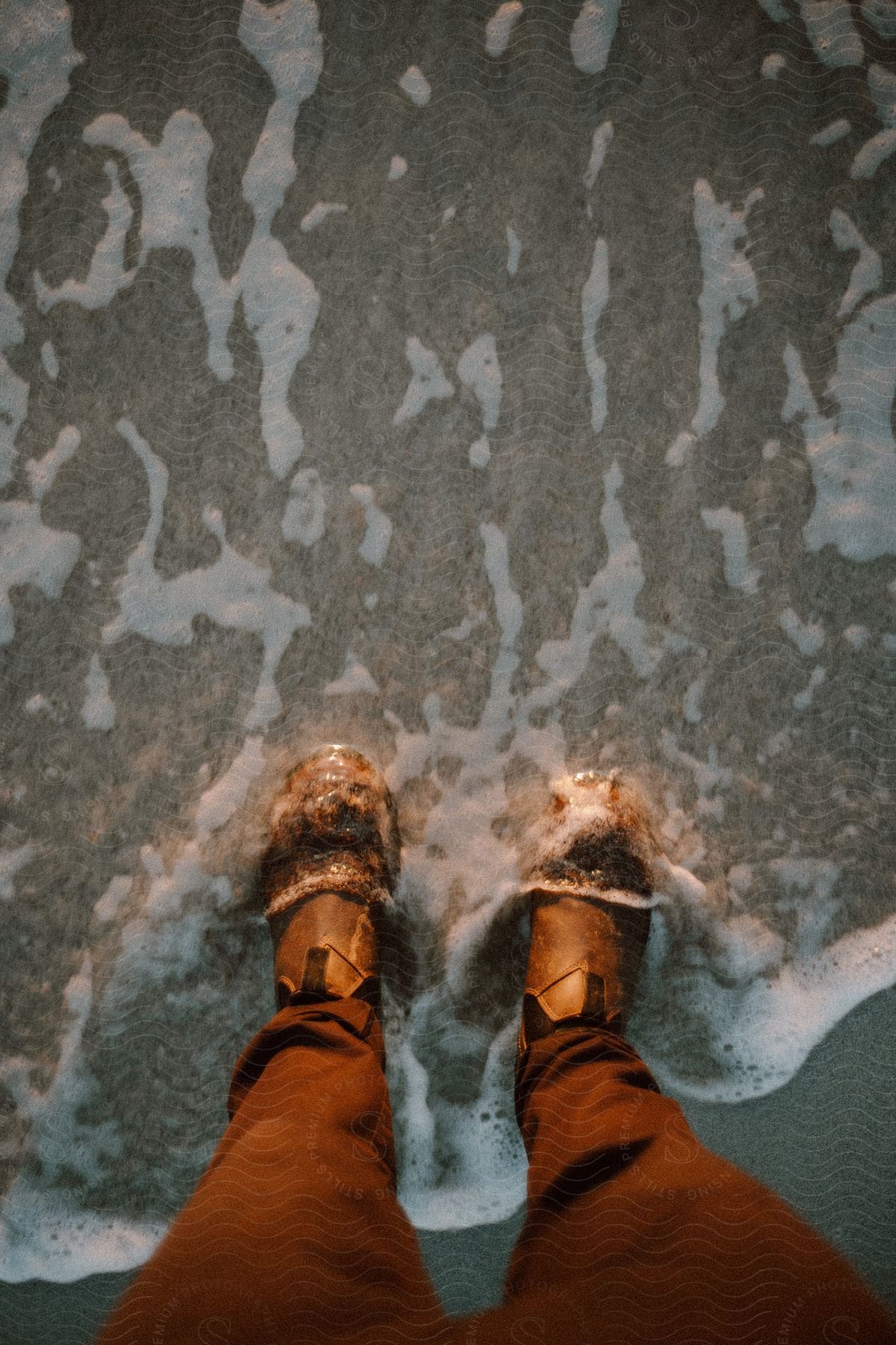 A person getting their feet wet on the beach but still wearing their shoes and pants.