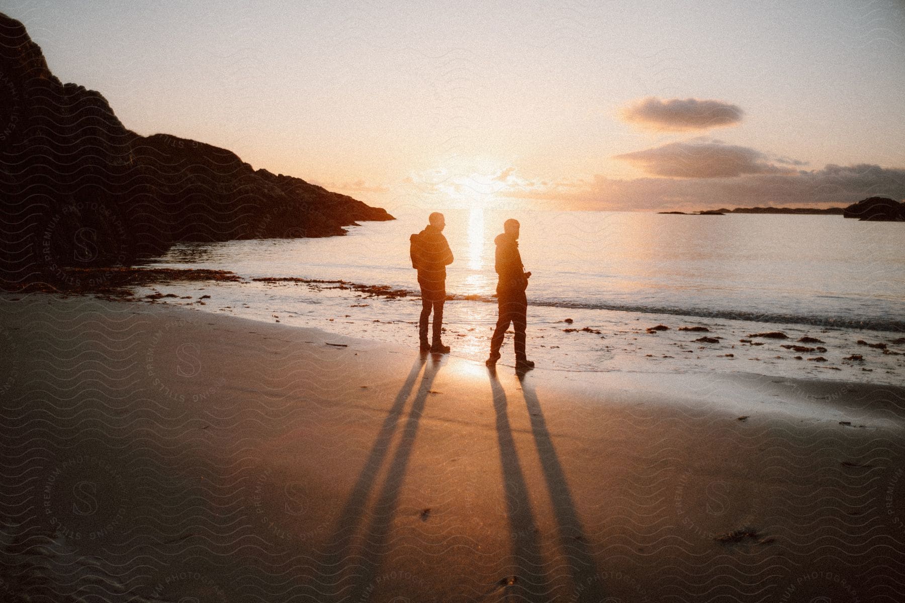 The silhouette of two people on the beach looking at the sea and the sun is shining brightly on the horizon