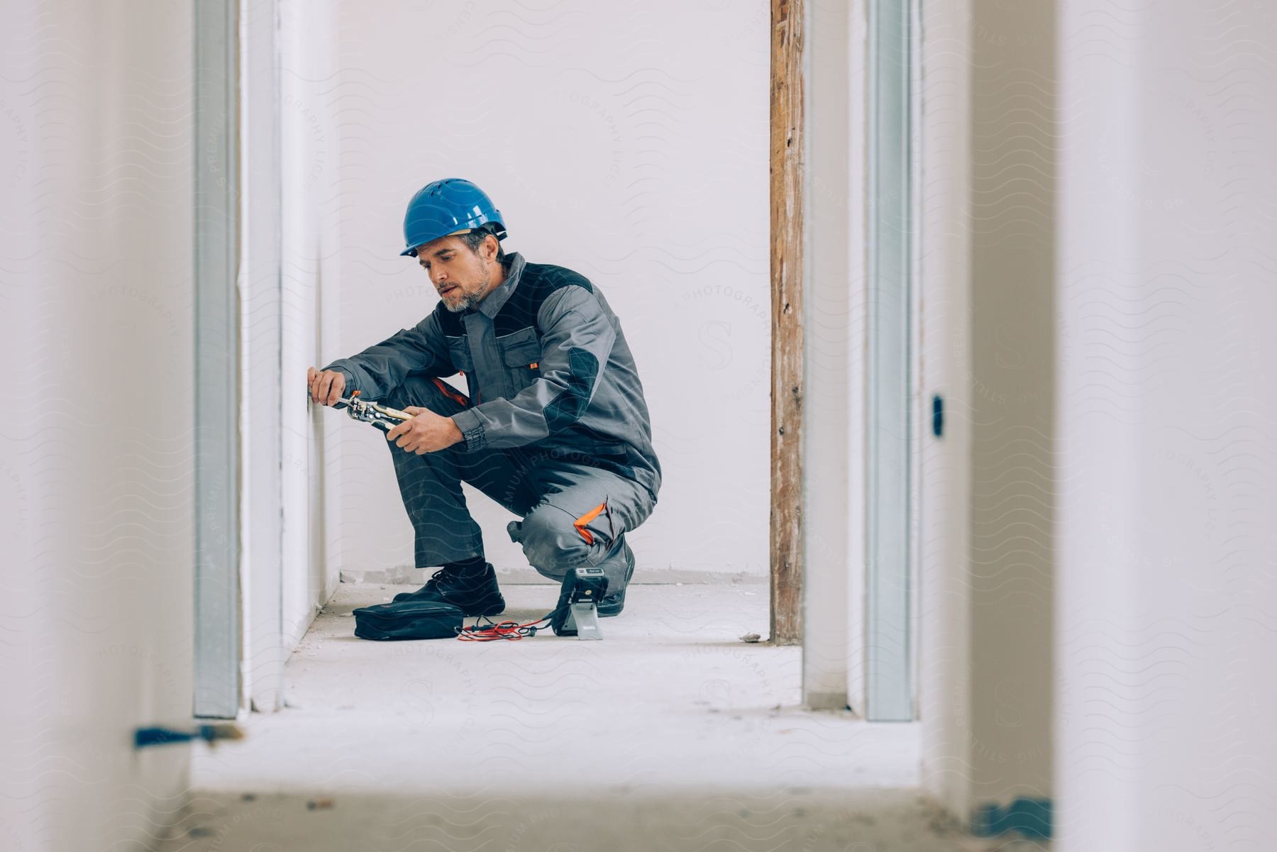 Construction worker drilling a hole in a wall, with a multimeter on the floor next to him.