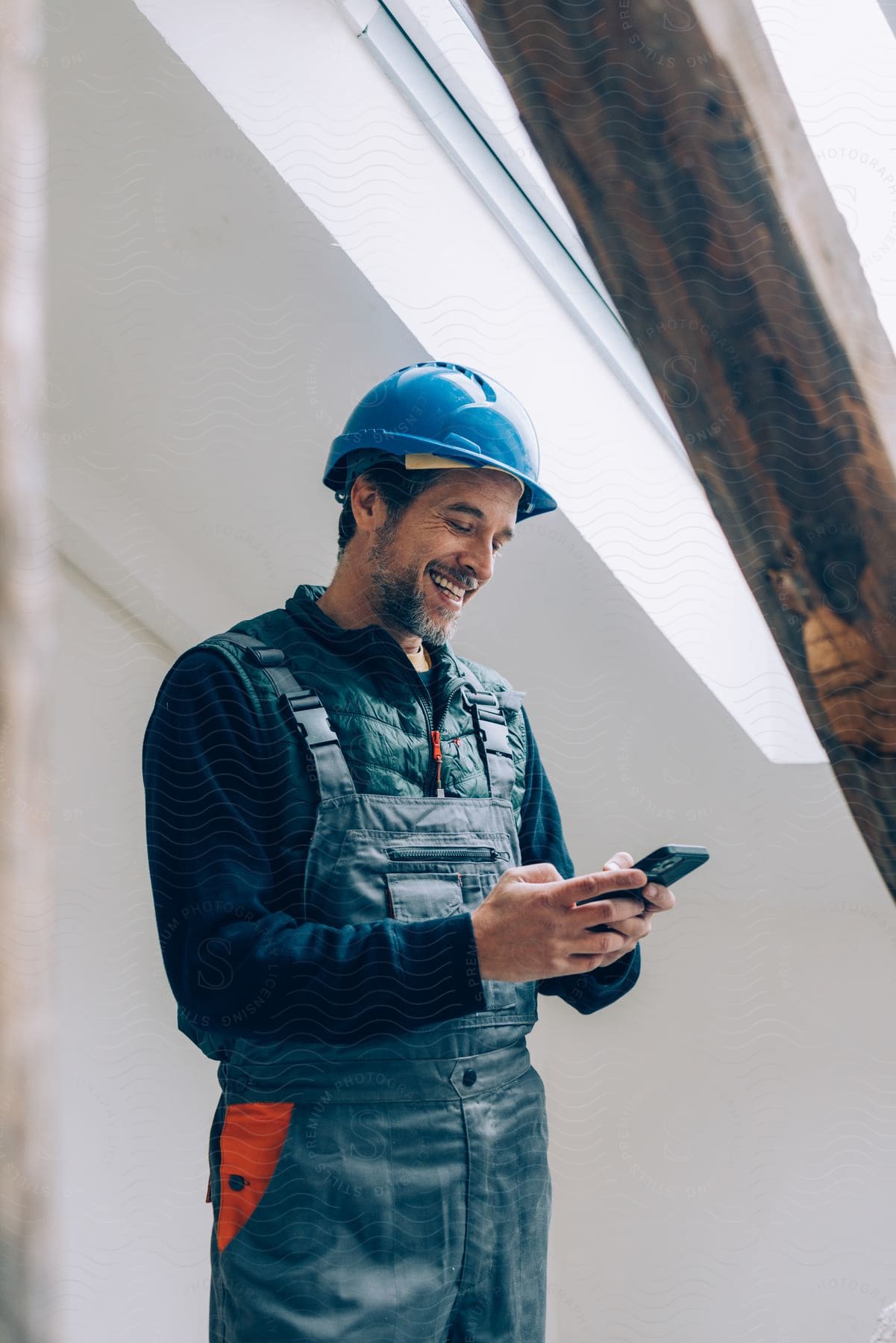 A construction worker smiling down at the phone in his hands standing near a wooden beam and a skylight